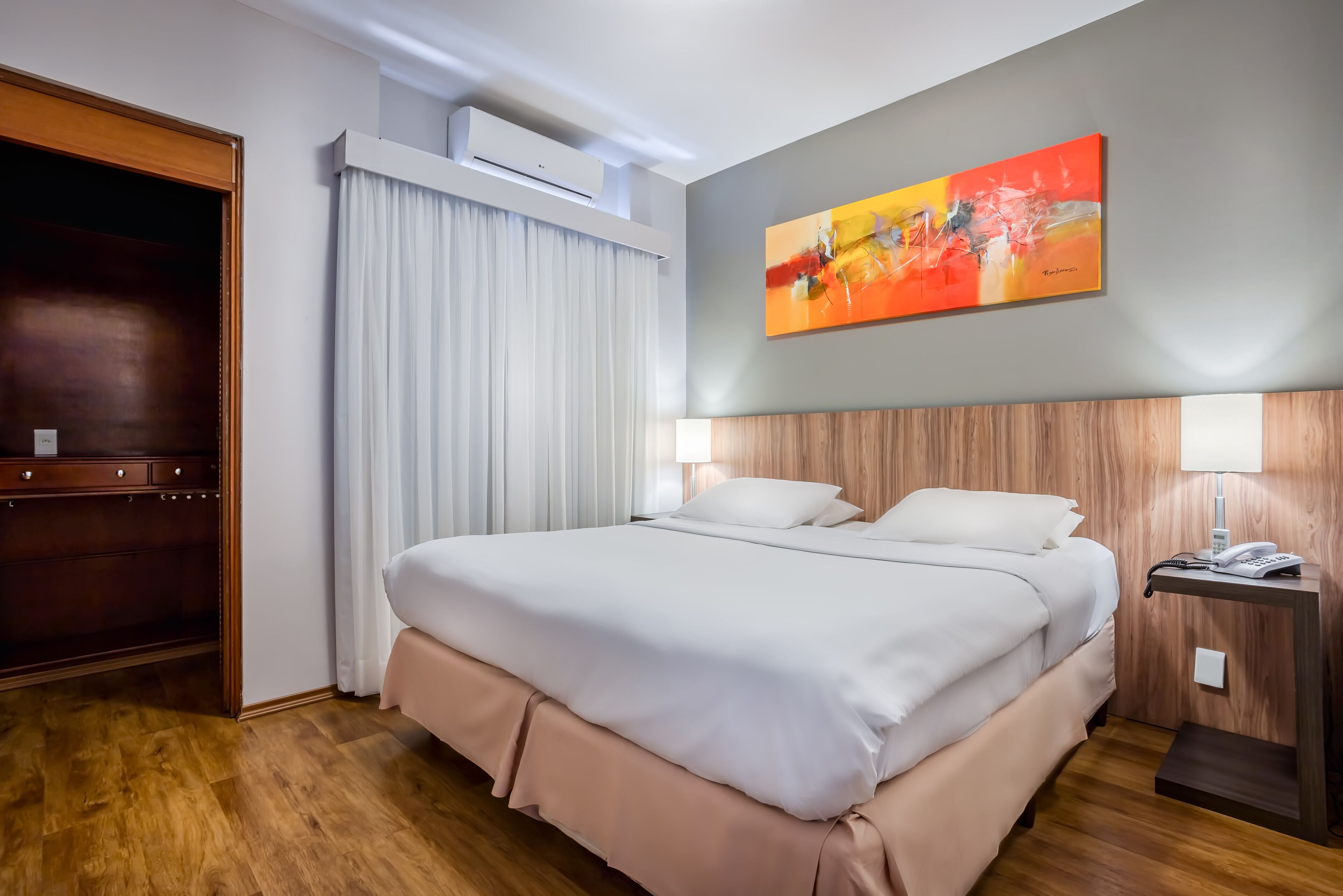 A hotel room with a large bed, a wooden headboard, a painting on the wall, a bedside table with a lamp, and a wardrobe.