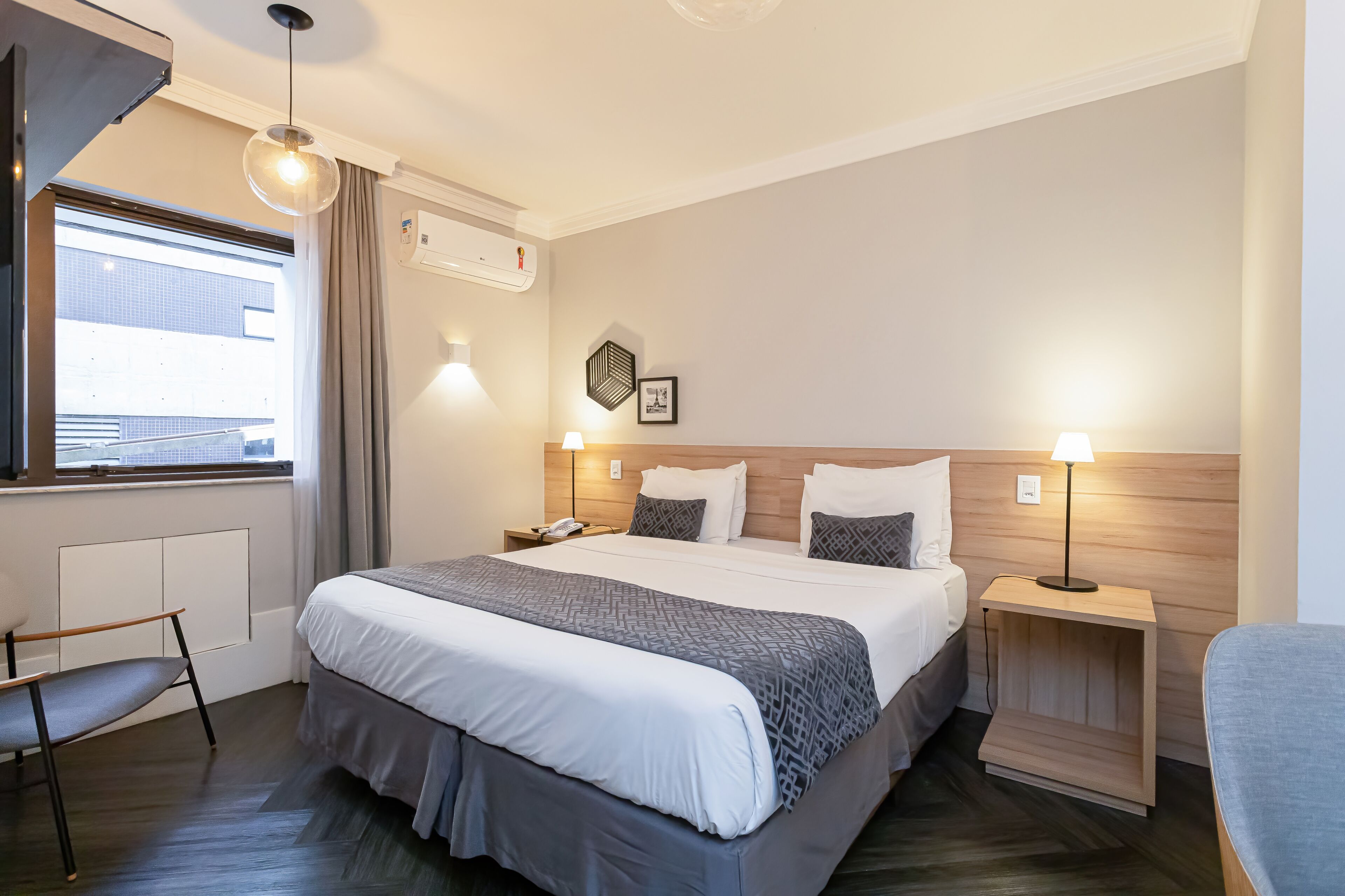 Une chambre d’hôtel avec un grand lit, des tables de chevet, une chaise, une fenêtre avec des rideaux et un climatiseur fixé au mur.
