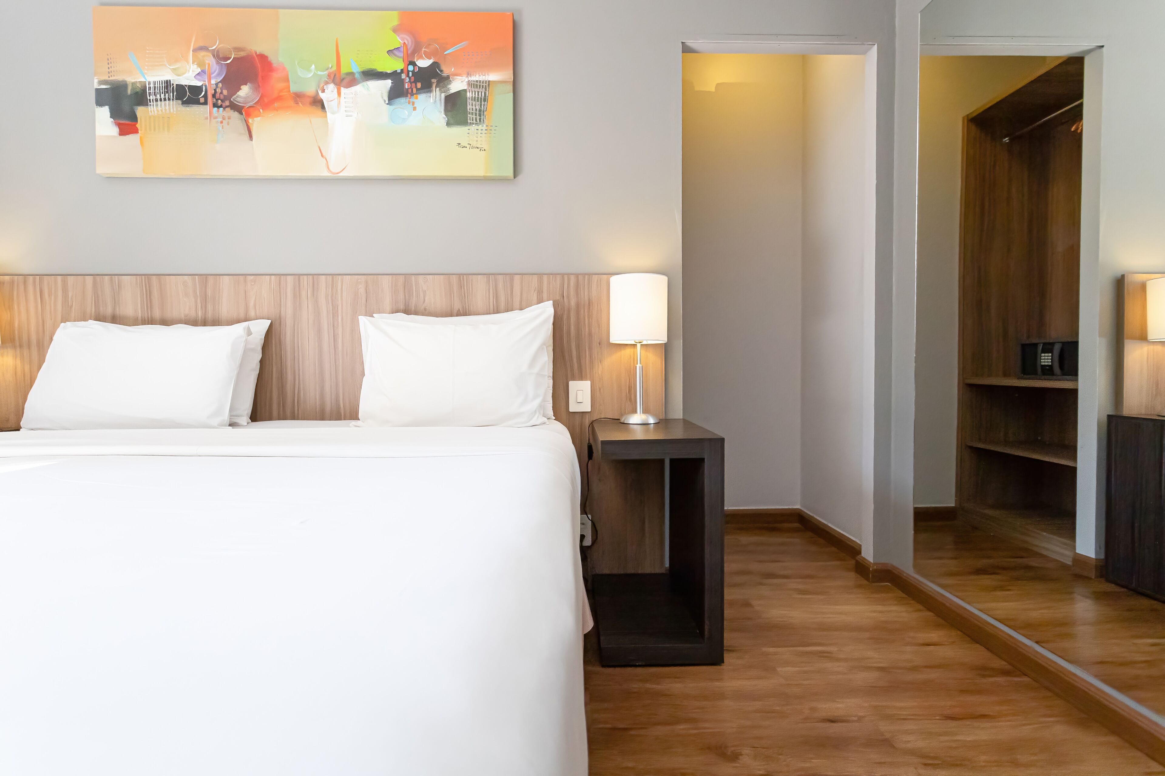 A hotel room with a large bed, a bedside table with a lamp, a wooden headboard, and a painting on the wall.