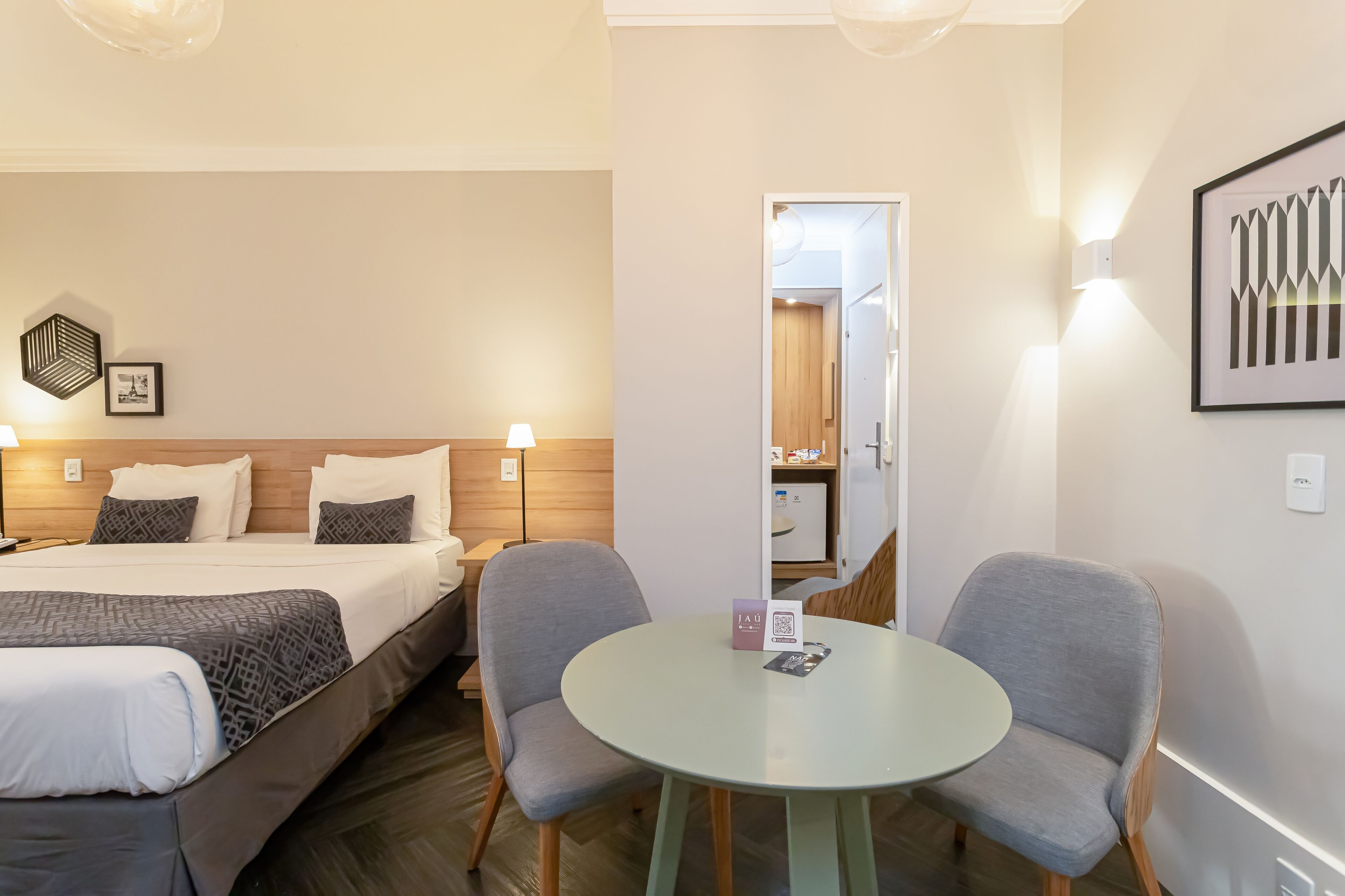 Ein Hotelzimmer mit einem Bett, zwei Stühlen, einem kleinen Tisch und einem Spiegel, der eine Tür und eine Küchenzeile zeigt.