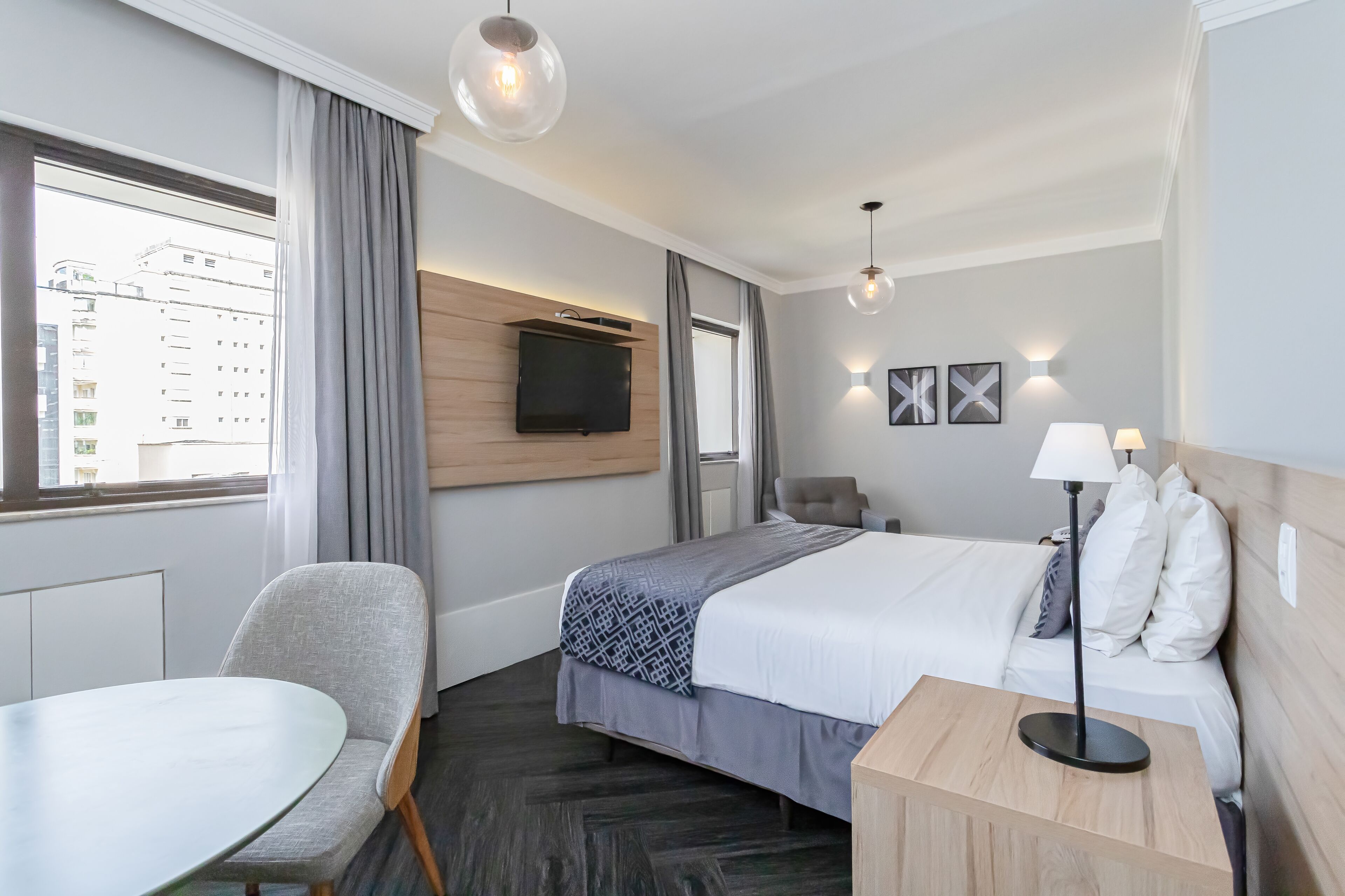 A hotel room with a bed, a desk, a chair, a TV mounted on the wall, and a window with curtains.