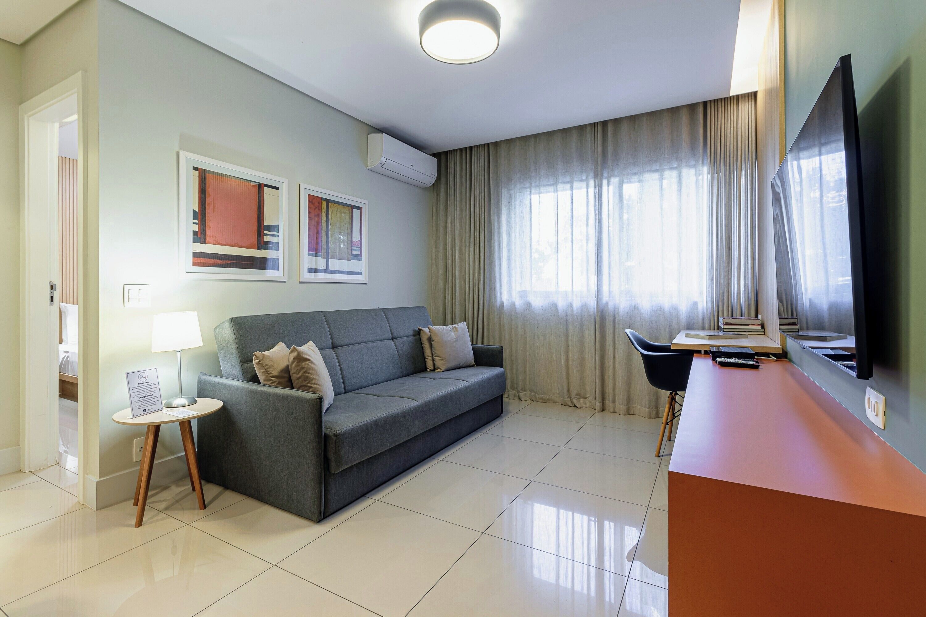 Een moderne woonkamer met een grijze bank, een klein rond tafeltje met een lamp, een flatscreen televisie en een groot raam met gordijnen.