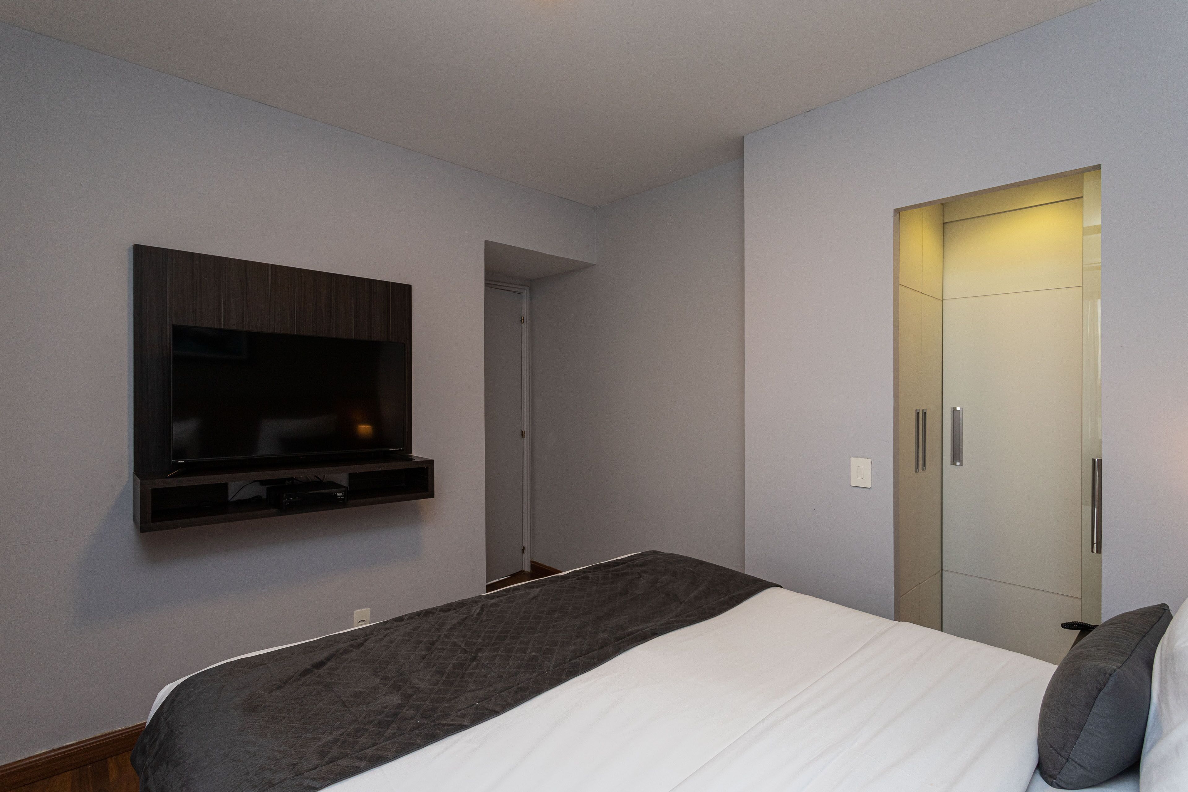 A modern hotel room with a large bed, a flat-screen TV mounted on the wall, and a wardrobe with a mirror.