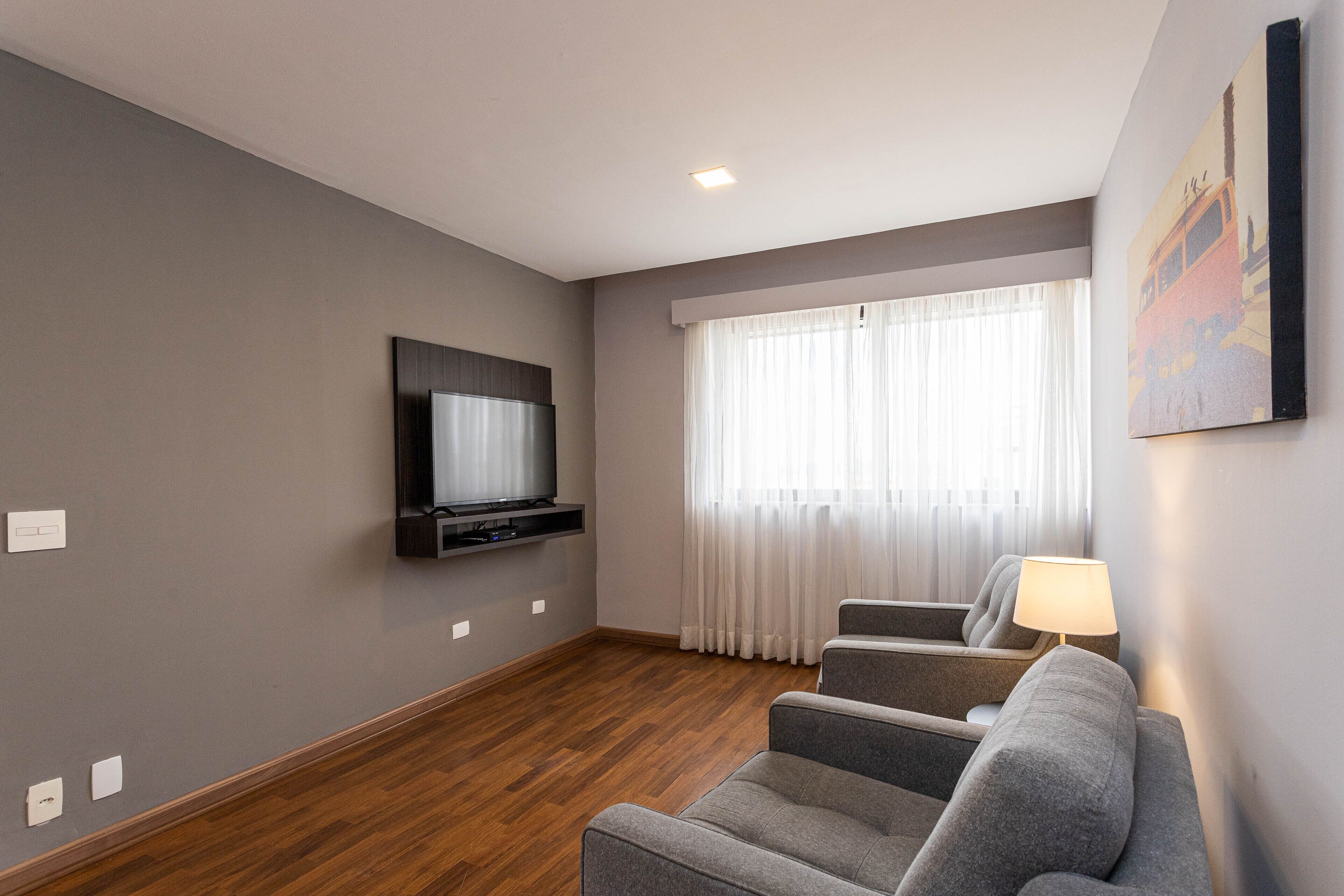 Een moderne woonkamer met een flatscreen televisie aan de muur, een grijze bank en een raam met dunne gordijnen.