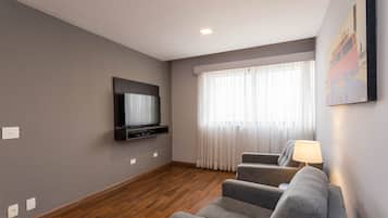 Uma sala de estar moderna com televisão de tela plana na parede, um sofá cinza e uma janela com cortinas leves.