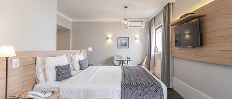Een moderne hotelkamer met een net opgemaakt bed, een nachtkastje met een lamp, een kleine tafel met een stoel, een aan de muur bevestigde televisie en een raam met gordijnen.