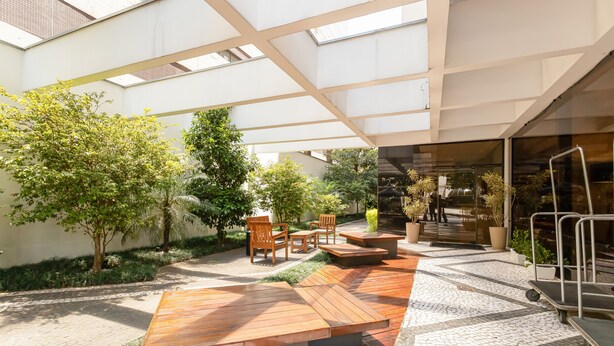 An outdoor patio area with wooden benches, a table, and chairs. There are potted plants and a pathway leading to a glass door. - H4 La Residence Paulista (São Paulo)