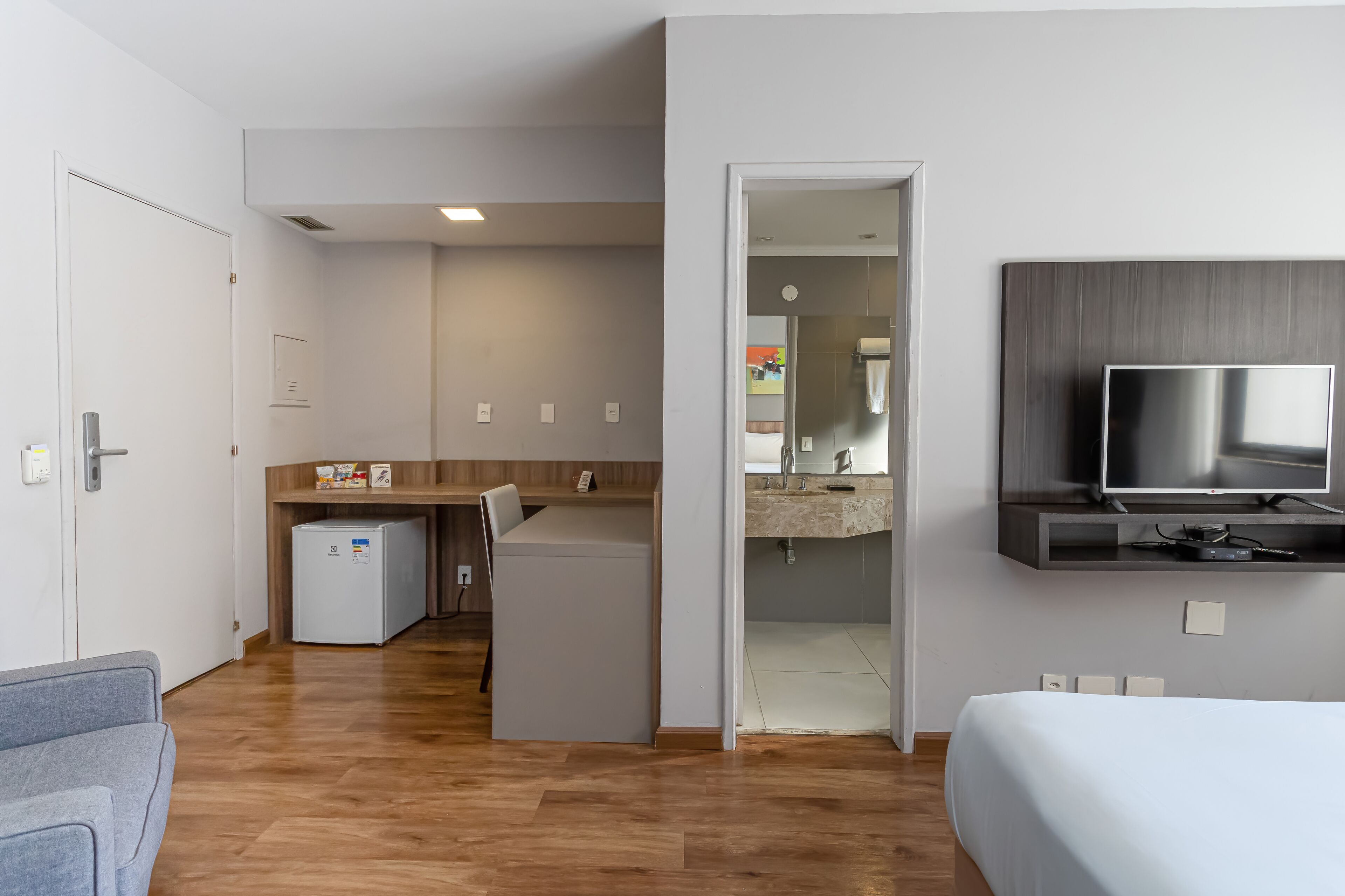 Habitación de hotel moderna con cama, televisor, una pequeña kitchenette y un baño visible a través de una puerta abierta.