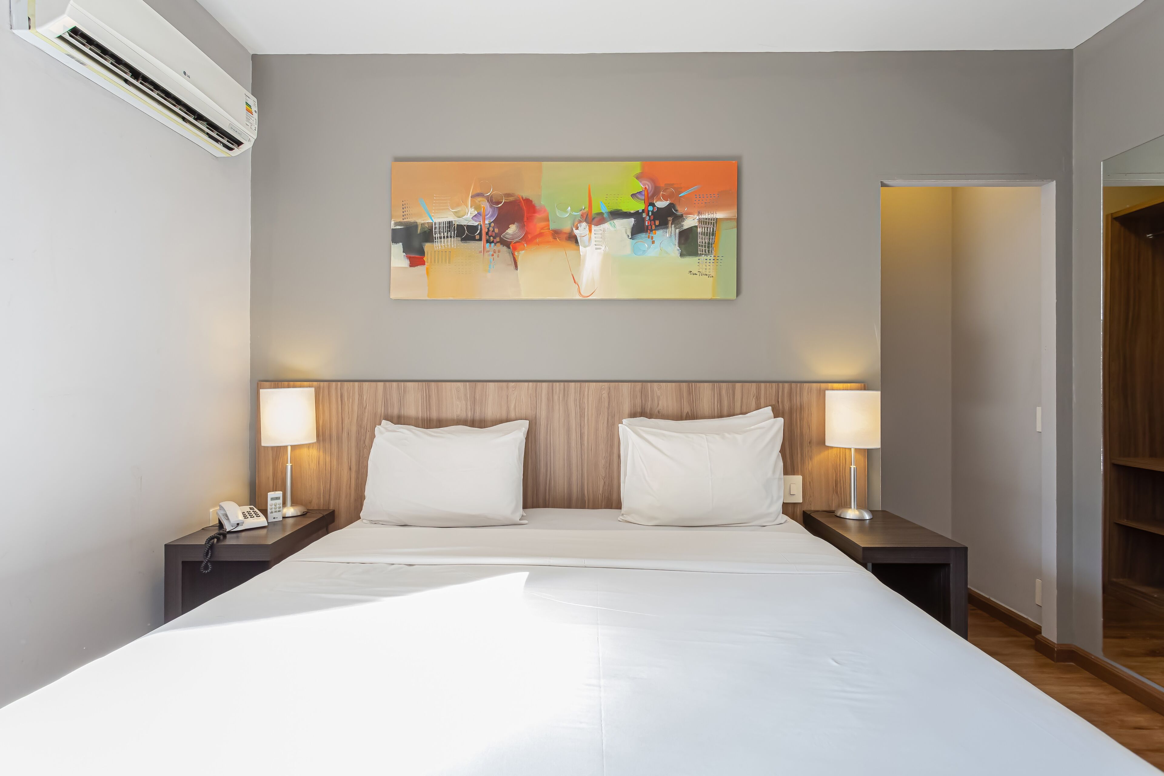 Ein modernes Hotelzimmer mit einem großen Bett, zwei Nachttischlampen, einem Bild an der Wand und einer Klimaanlage.