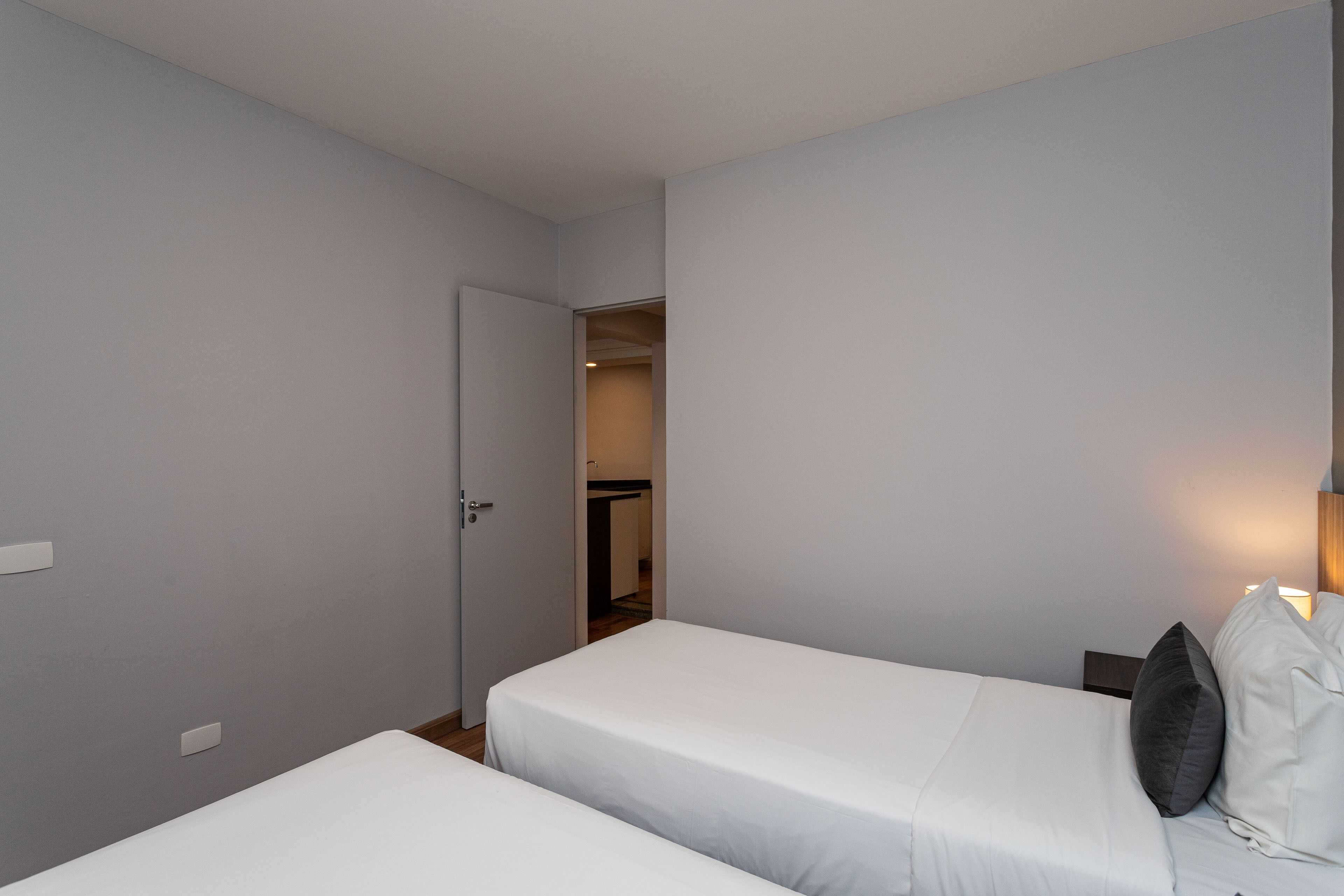 A small, clean room with two single beds, a door leading to a bathroom, and a nightstand with a lamp.
