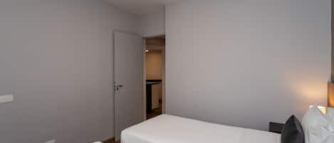 A small, clean room with two single beds, a door leading to a bathroom, and a nightstand with a lamp.