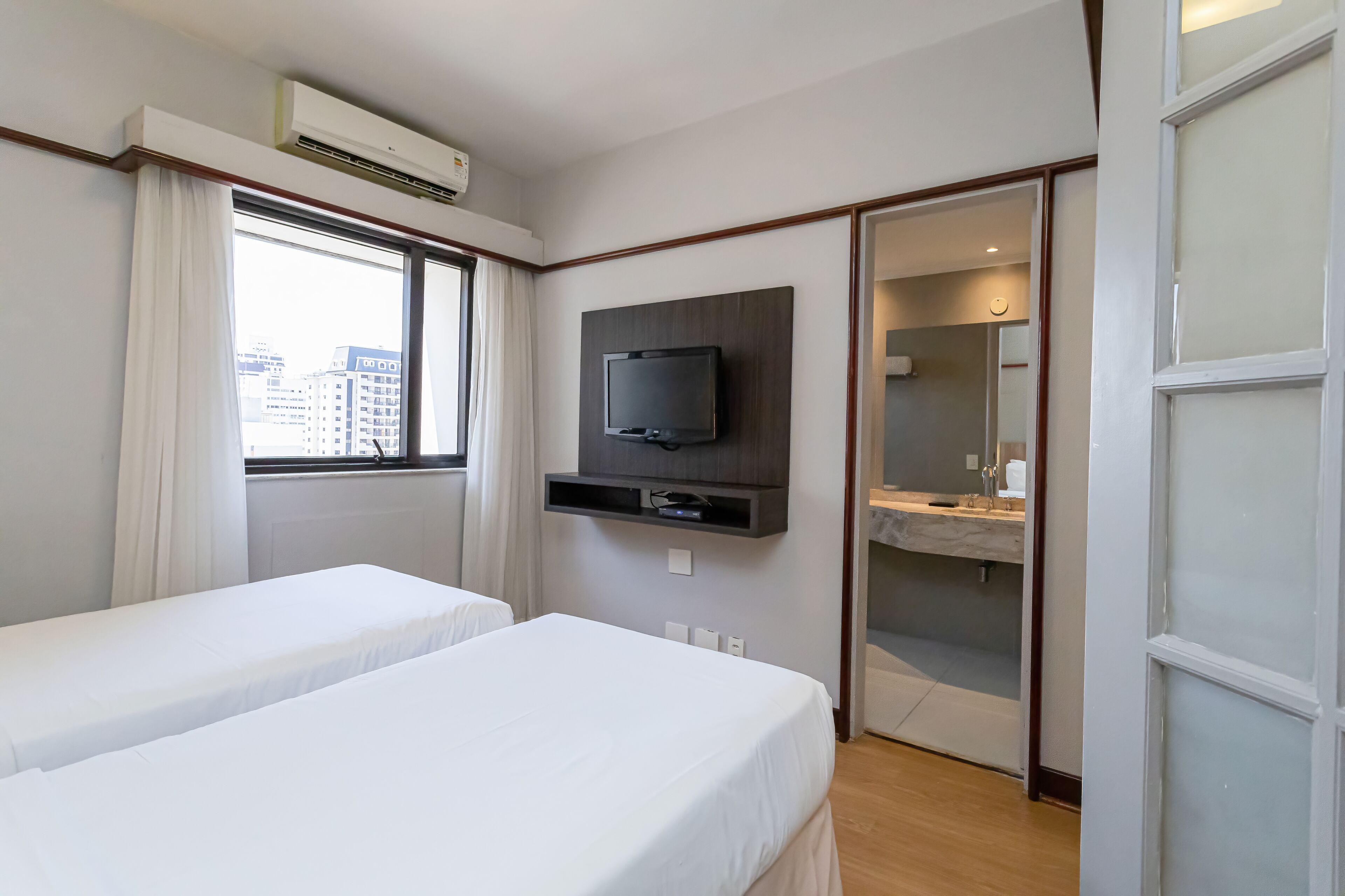 A hotel room with two single beds, a TV mounted on the wall, an air conditioning unit above the window, and a bathroom visible through a glass door.