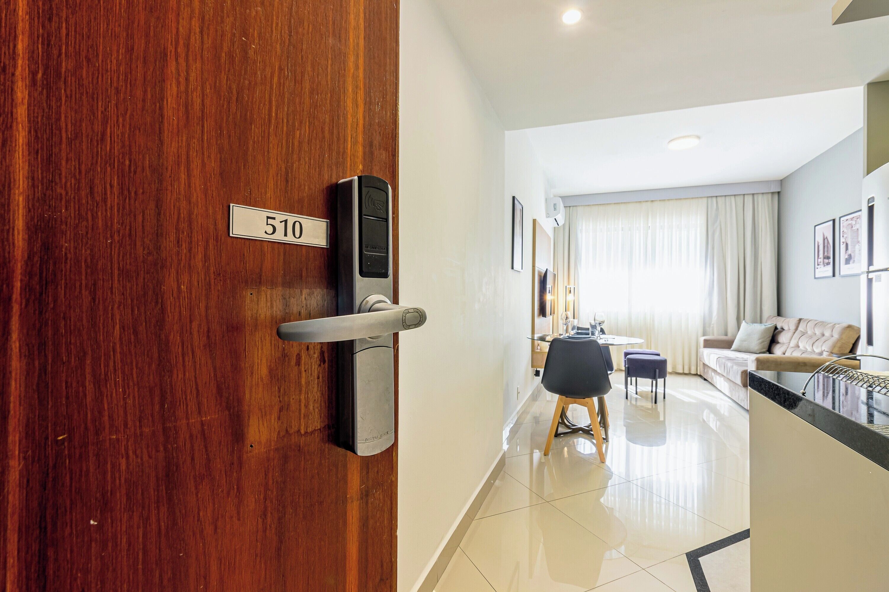 A hotel room with a wooden door, a digital lock, a dining area with a table and chairs, a sofa, and a large window with curtains.