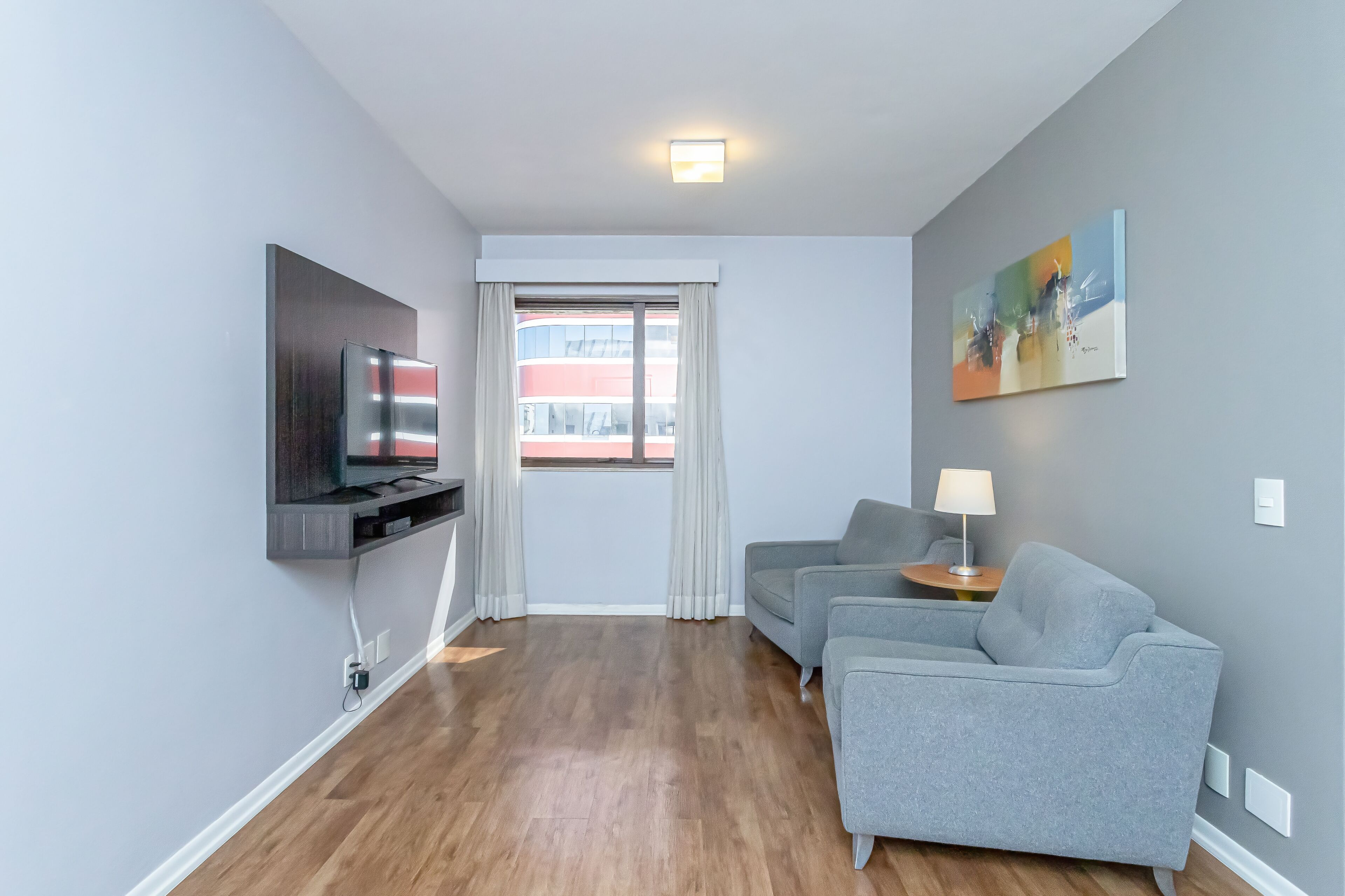 A modern living room with a flat-screen TV, grey sofas, a small table with a lamp, and a large window with curtains.