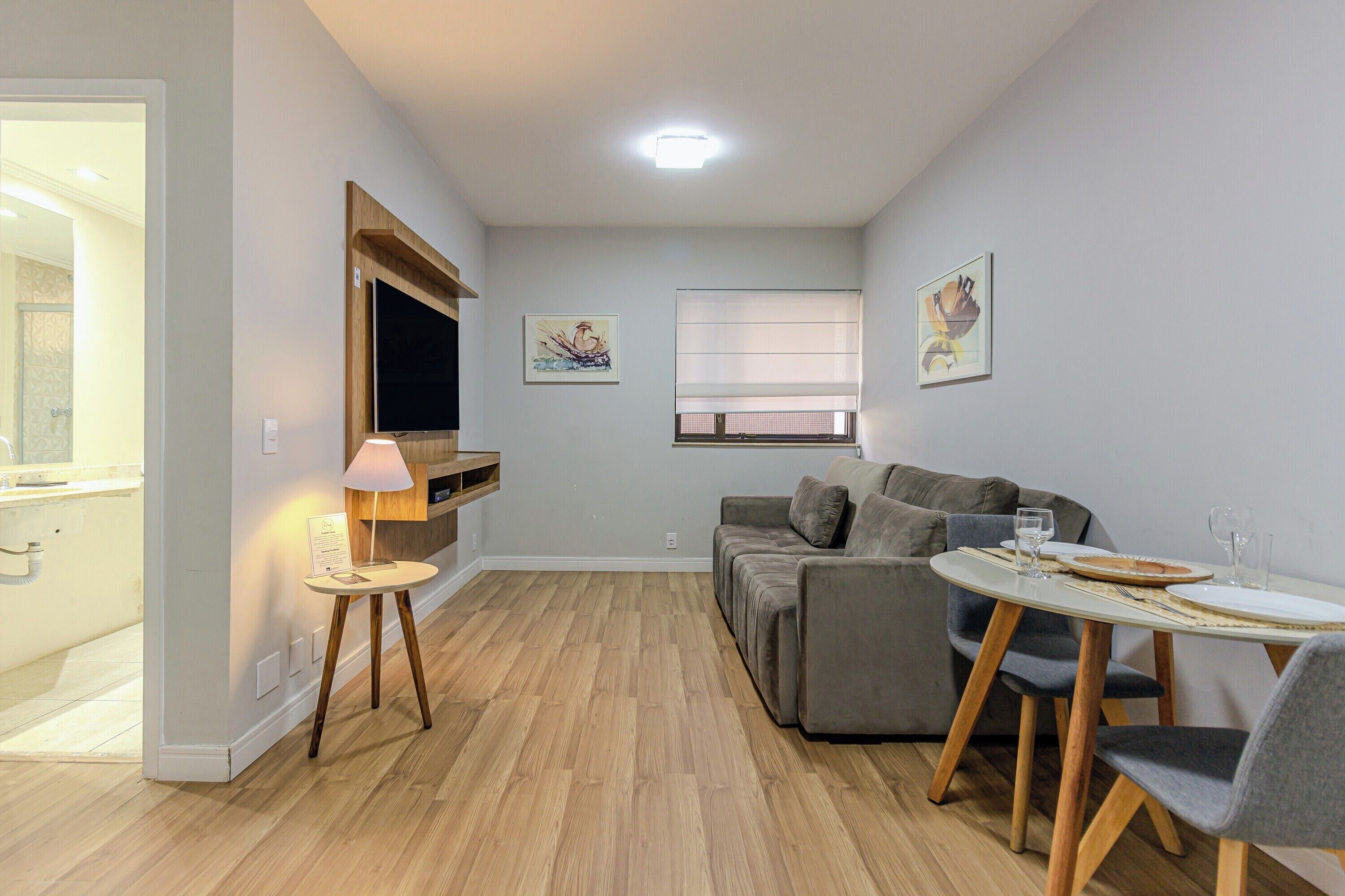 Un salon moderne avec un canapé gris, une table à manger avec des chaises, un téléviseur fixé au mur et des œuvres d’art accrochées aux murs.