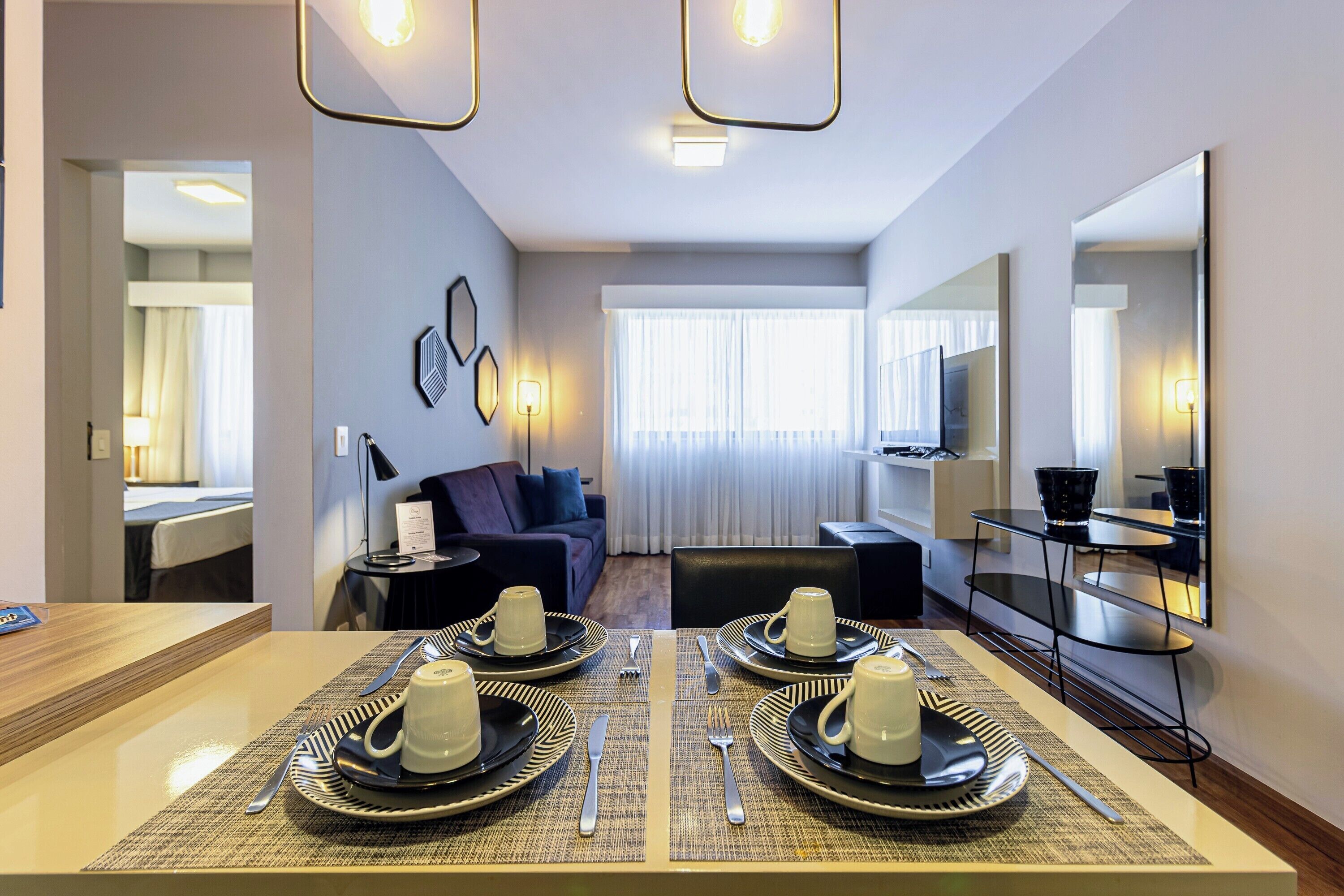 Une chambre d’hôtel moderne avec un coin repas aménagé pour deux personnes, un canapé, une télévision et un grand miroir.