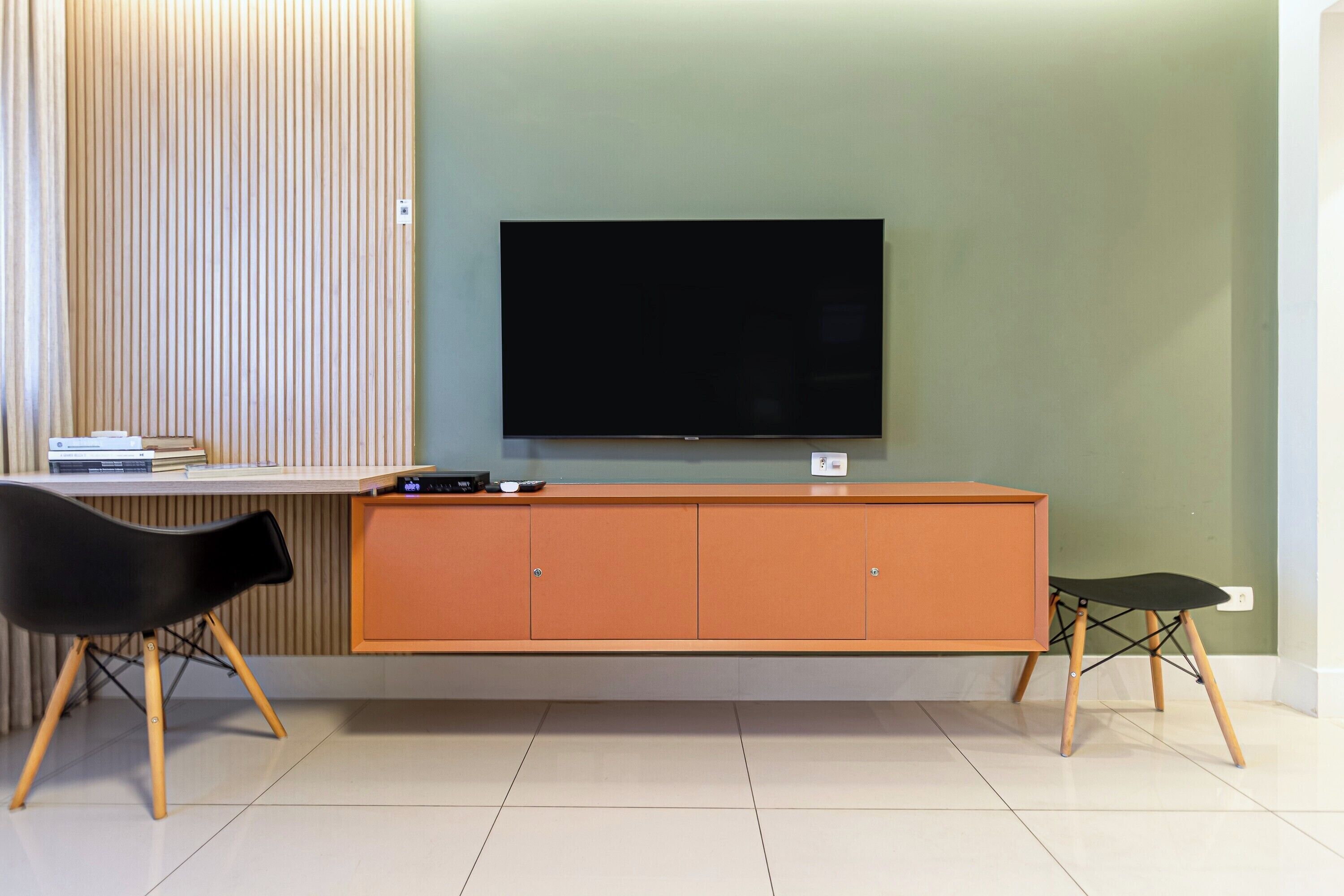 Ein modernes Wohnzimmer mit Flachbildfernseher an der Wand, einem Holzschrank mit TV-Stand, zwei schwarzen Sesseln und einem kleinen Tisch mit Büchern und elektronischen Geräten.