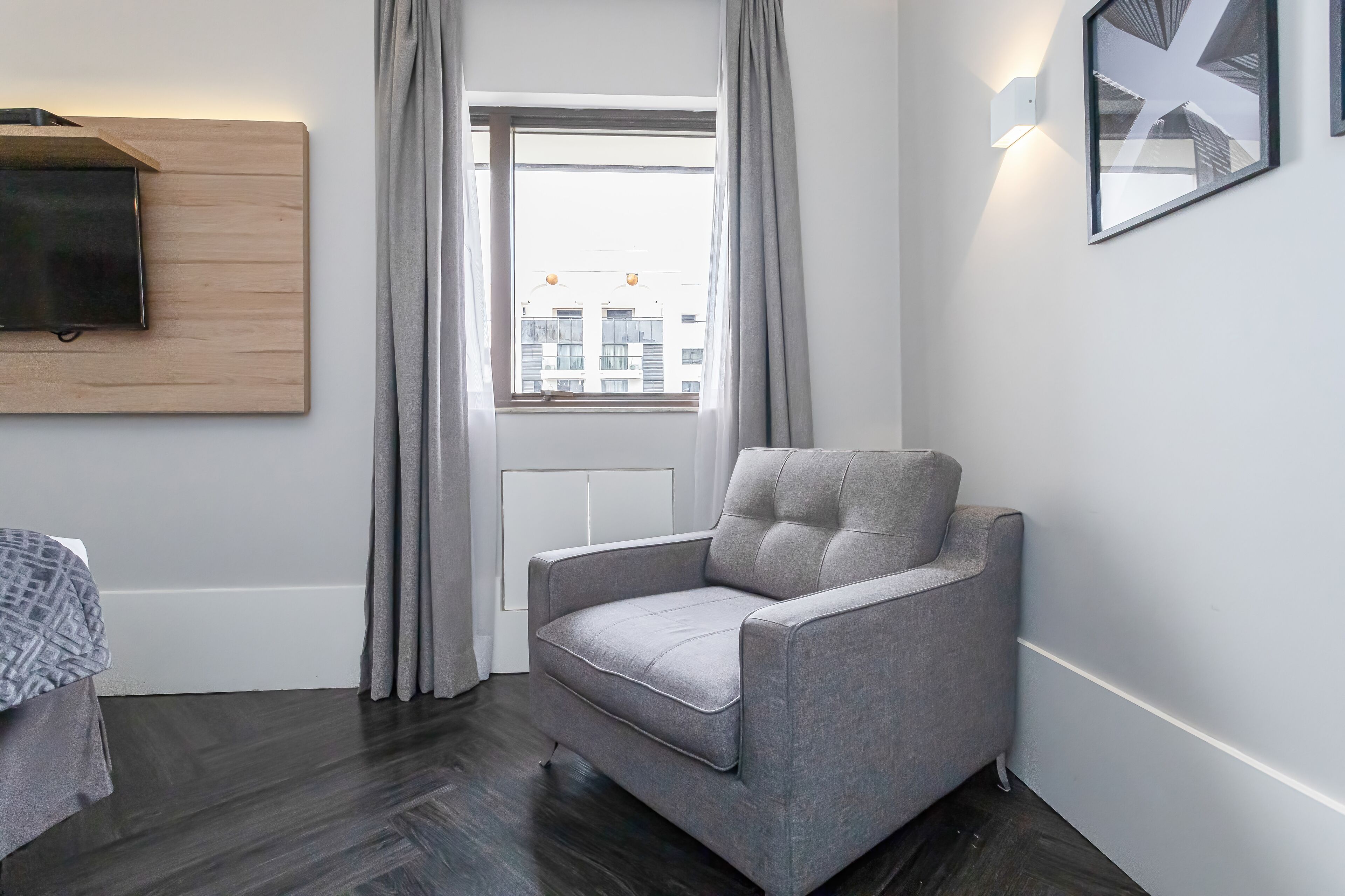 A modern room with a grey upholstered armchair, a wooden panel wall, a flat-screen TV, and a window with curtains.