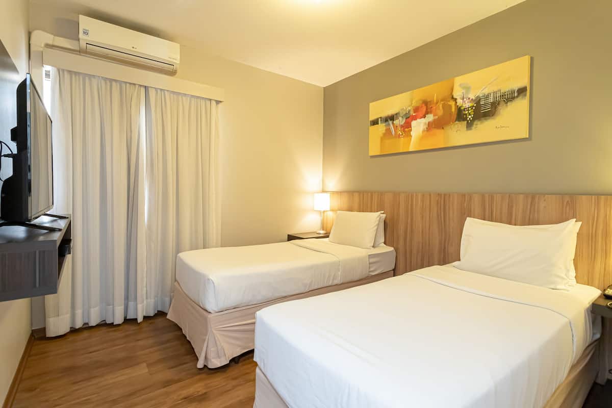 A hotel room with two single beds, a TV, a painting on the wall, and an air conditioning unit.