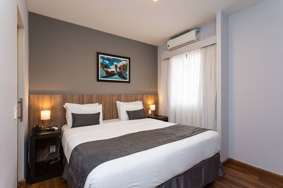 A hotel room with a large bed, two bedside tables with lamps, a painting on the wall, and an air conditioning unit above the window.