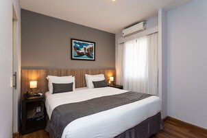 Una camera d'albergo con un letto grande, due comodini con lampade, un quadro al muro e un condizionatore d'aria sopra la finestra.