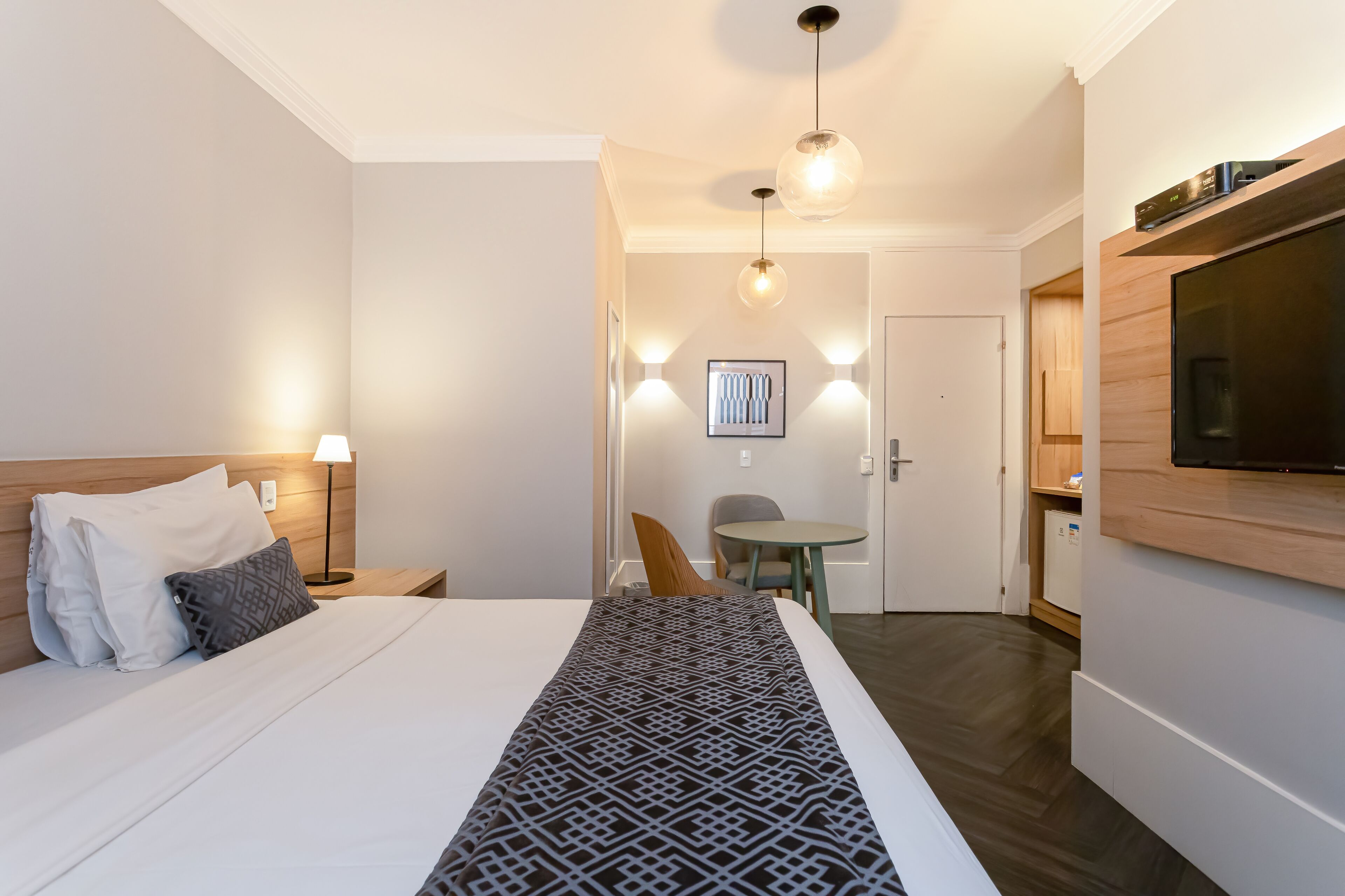 Habitación de hotel con cama, lámparas de noche, televisor montado en la pared, una mesita y una silla.