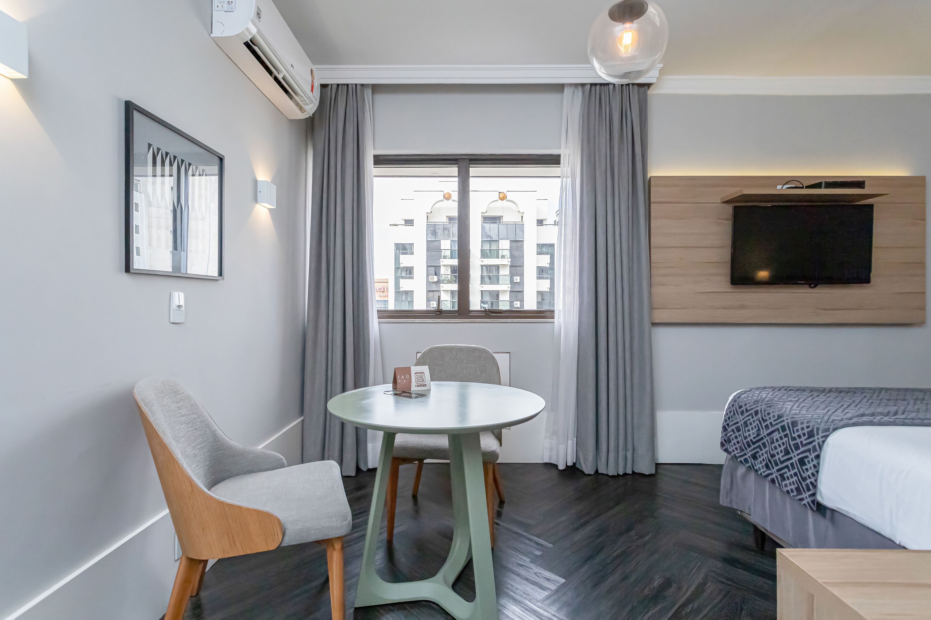 A modern hotel room with a dining table, chairs, a bed, a TV mounted on the wall, and a window with curtains.