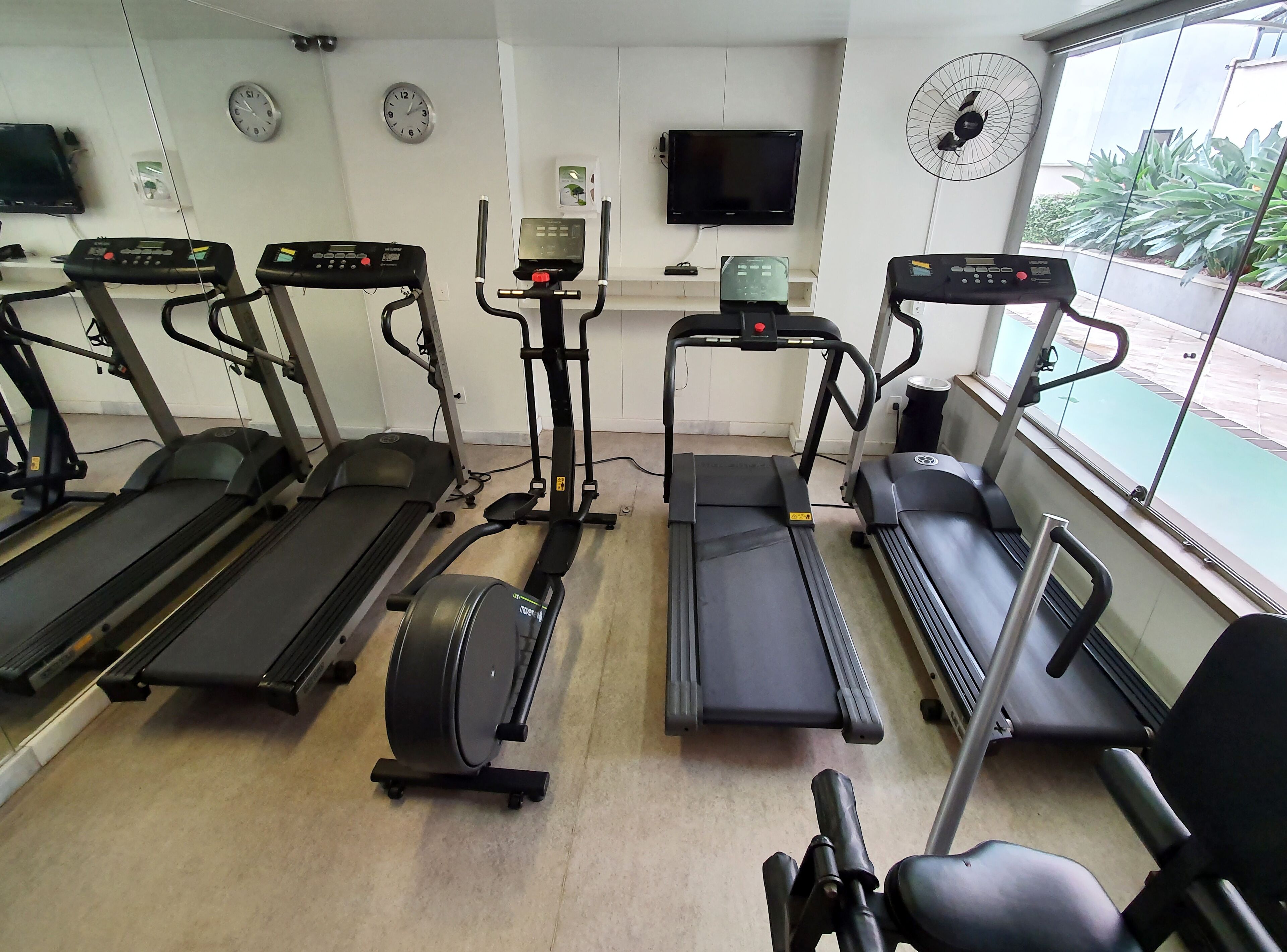 A gym with treadmills, elliptical machines, and a stationary bike. There are clocks on the wall, a TV, and a window with a view of the outside.