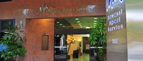 The entrance to Max Savassi Apart Service, featuring a modern design with a reception desk, potted plants, and a clear view into the interior.