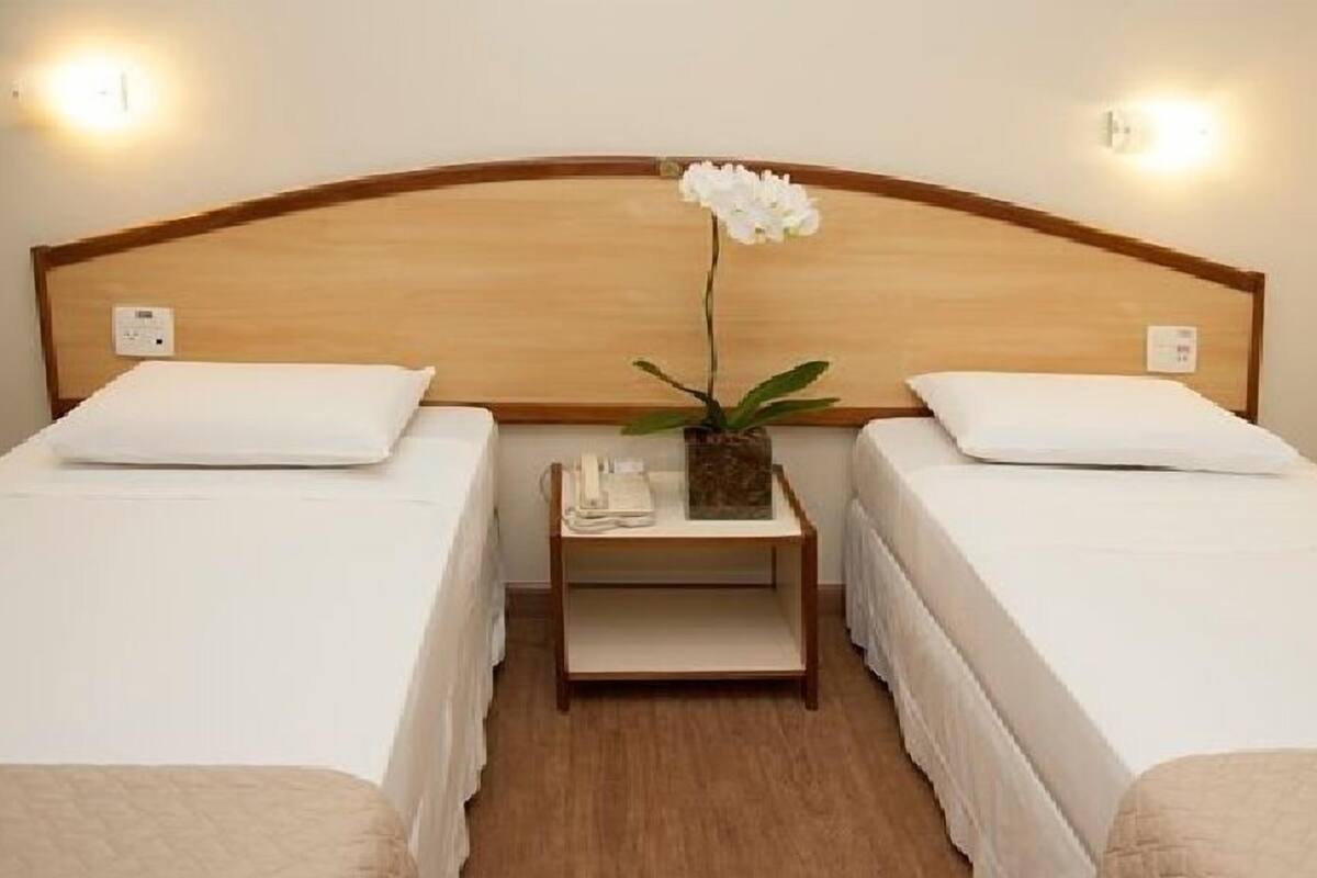 Une chambre d’hôtel avec deux lits simples, une tête de lit en bois, des lampes de chevet, une table de chevet avec un téléphone et une plante, et une petite table entre les lits.