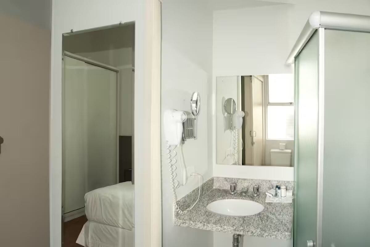 A hotel bathroom with a white sink, a mirror, a hairdryer, and a towel hanging on the wall.