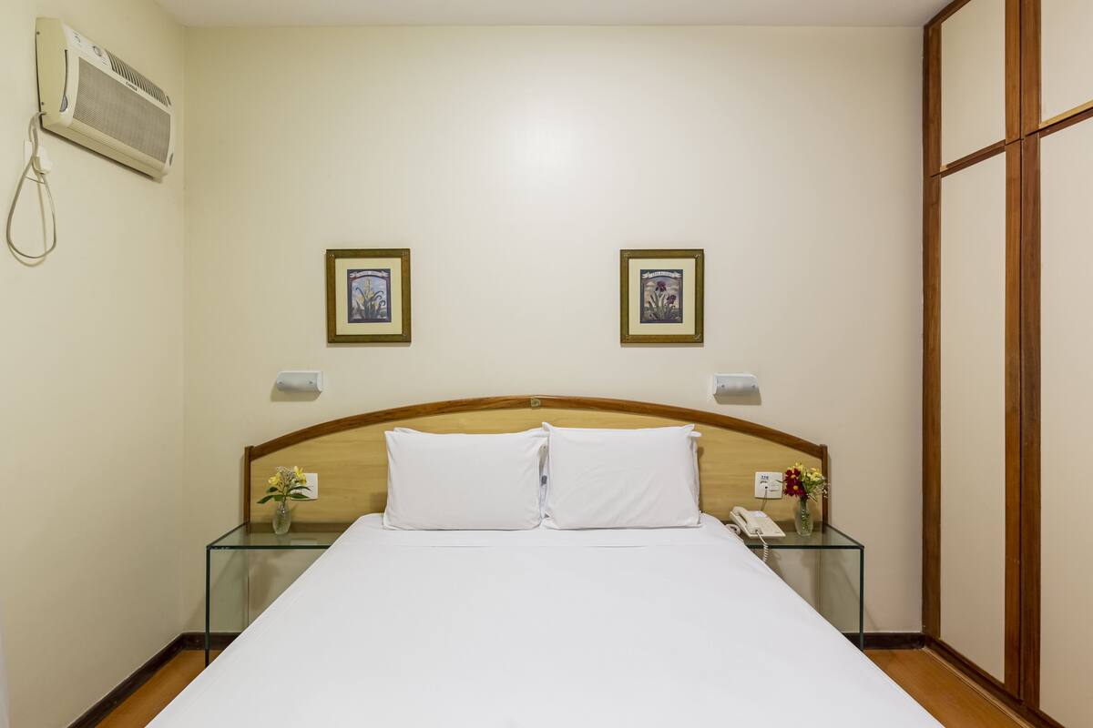 Une chambre d’hôtel comprenant un lit, deux tables de chevet, une petite table, un climatiseur fixé au mur et deux tableaux encadrés accrochés au mur.