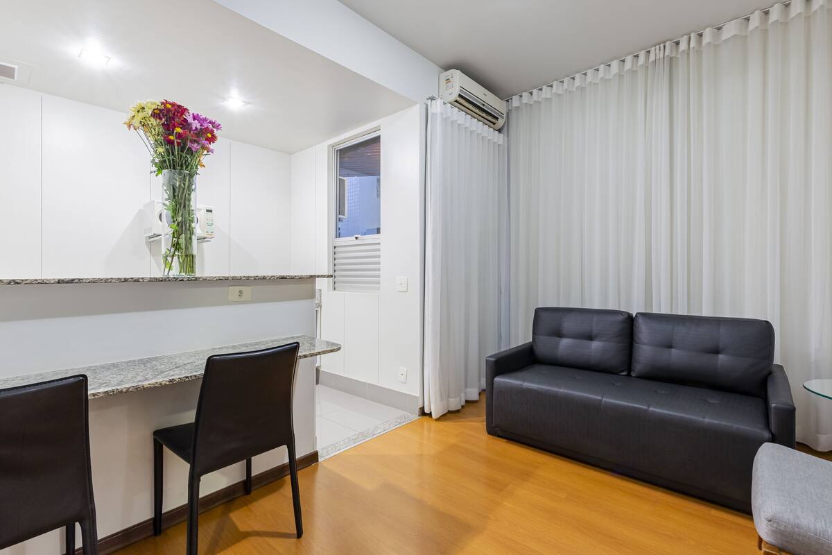 Un salon moderne avec un canapé noir, un coin repas comprenant une table et des chaises, et un espace cuisine avec un comptoir et un vase de fleurs.