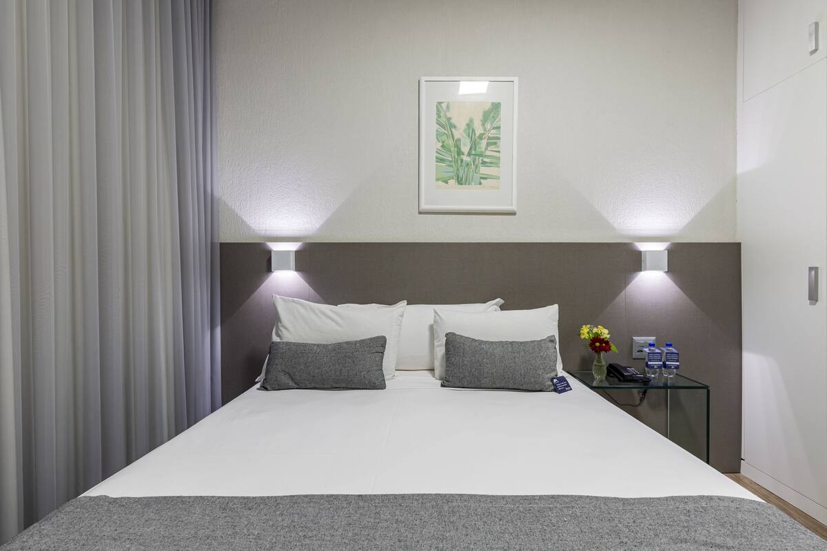 Une chambre d’hôtel avec un grand lit, deux lampes de chevet, une estampe botanique encadrée au mur, et une petite table sur laquelle se trouvent un vase de fleurs et un téléphone.