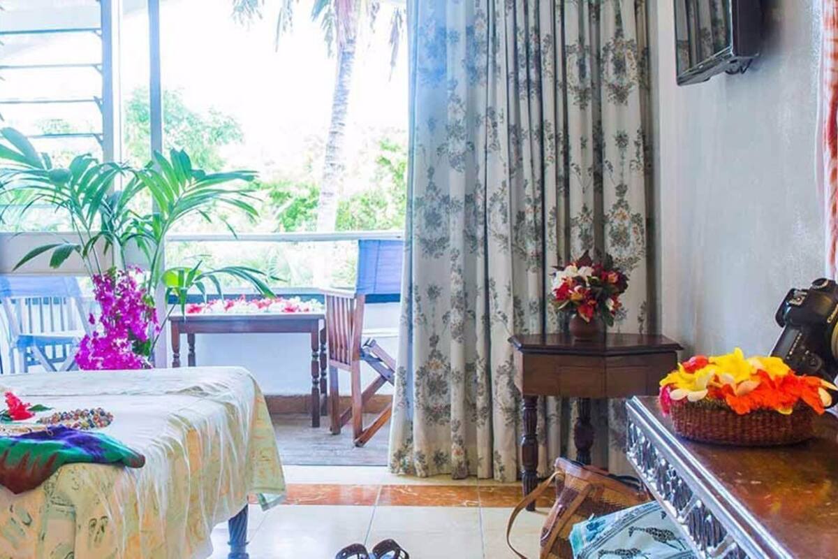 Habitación de hotel con cama, una mesita con una planta, un balcón con vistas y una cesta de mimbre con flores.