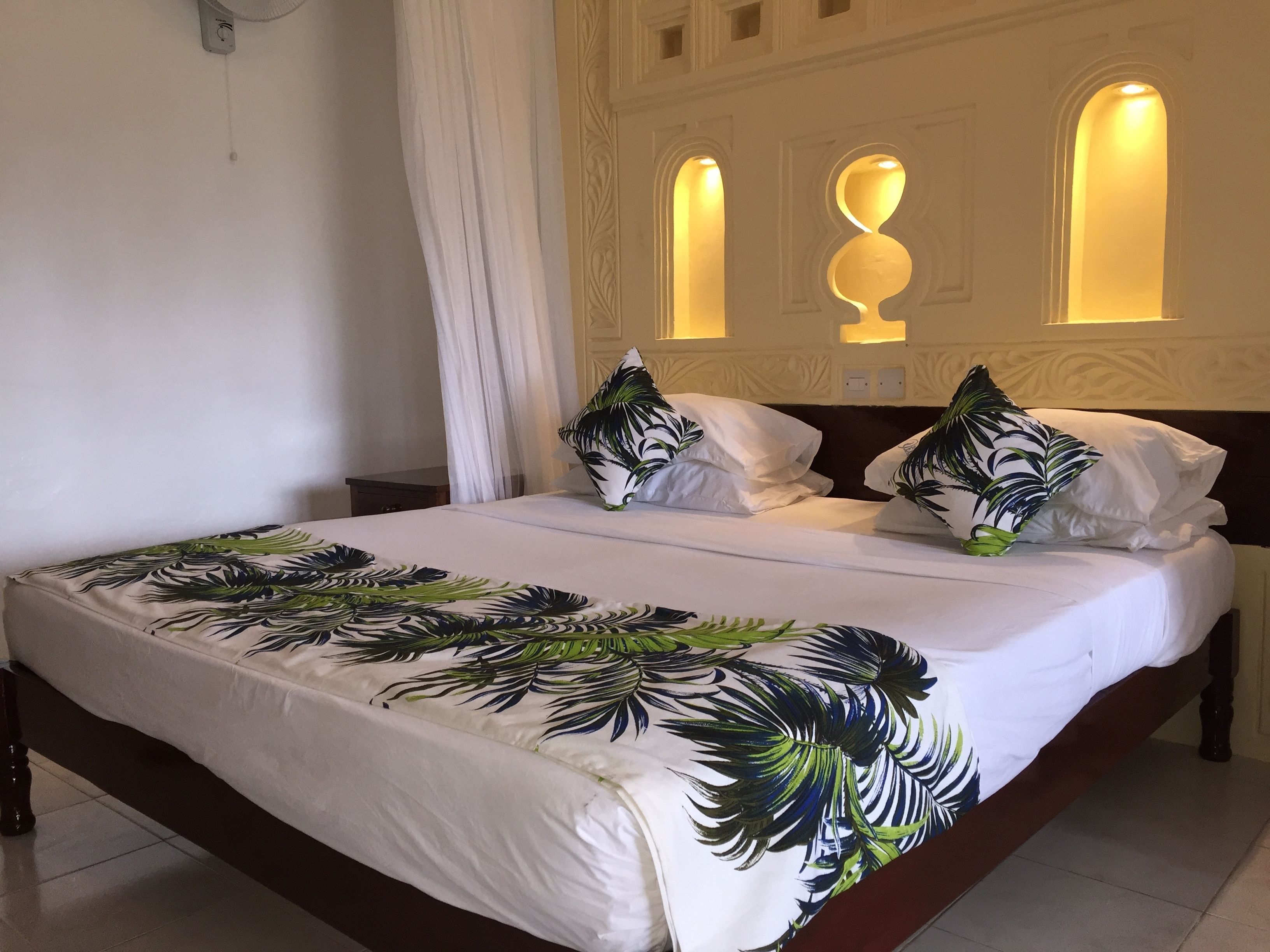 A neatly made bed with a tropical leaf-patterned bedspread, white pillows, and a wooden headboard.