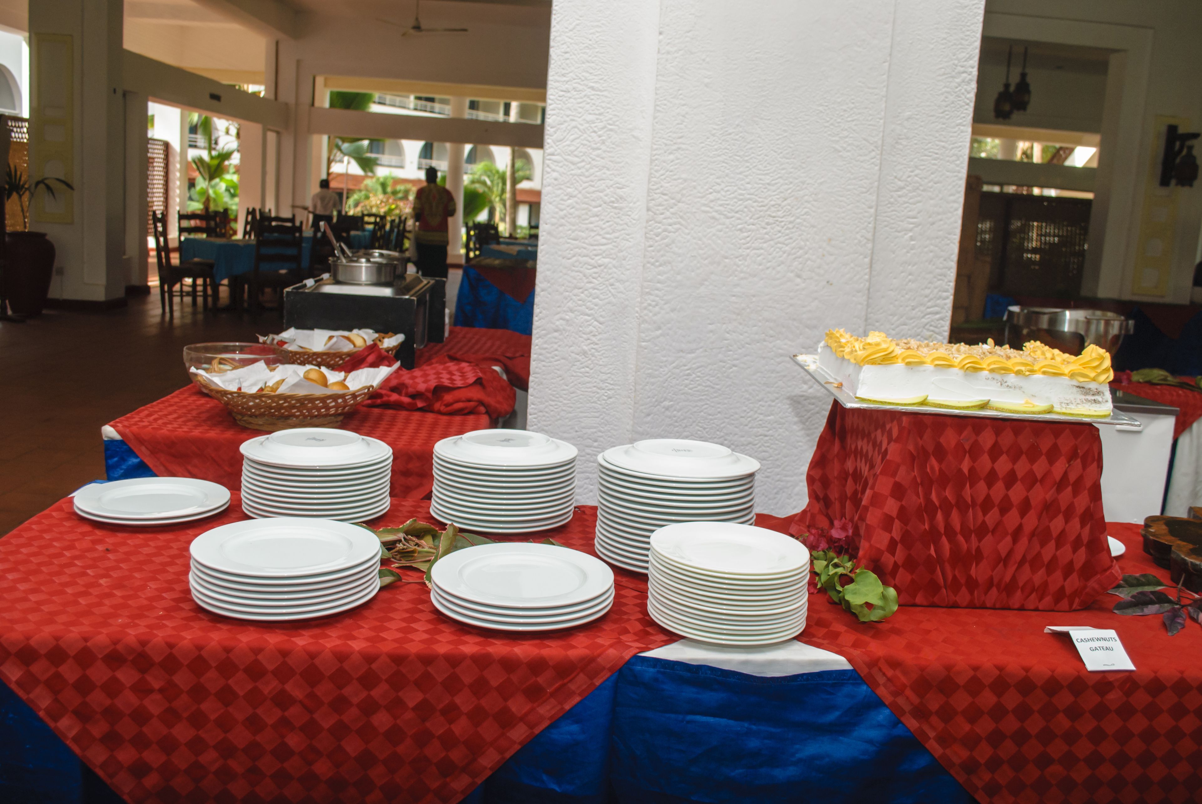 A buffet setup with multiple tables covered in red and blue tablecloths, featuring stacks of white plates and a tray of yellow food.