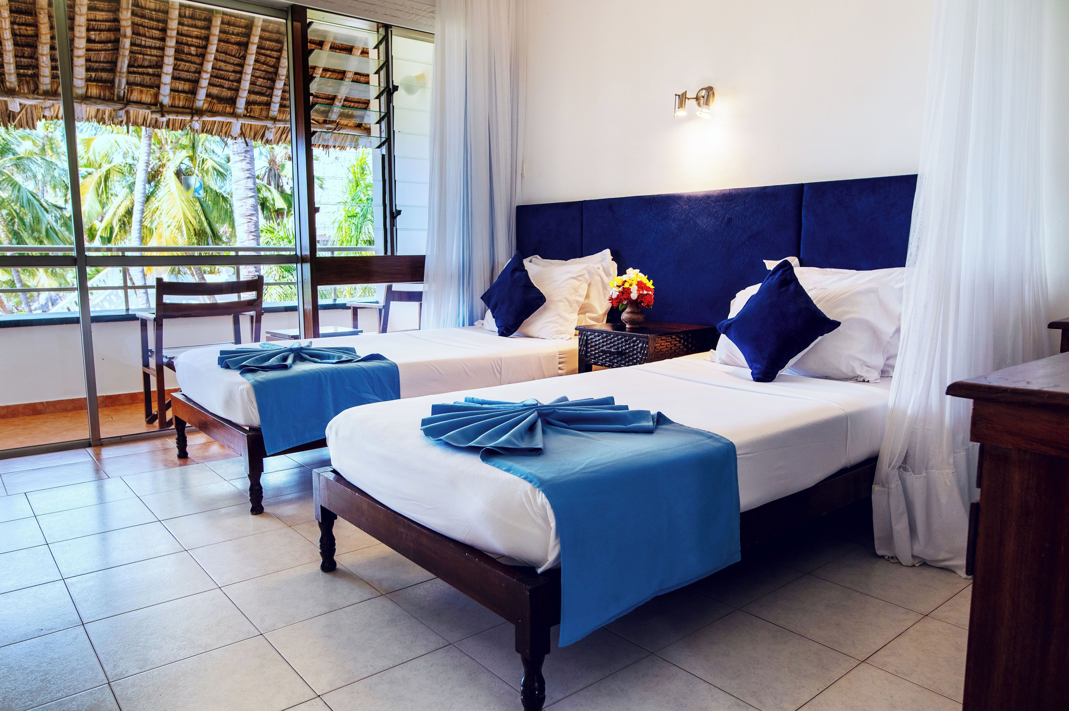 A hotel room with two beds, a wooden headboard, a window with a view of palm trees, and a chair by the window.