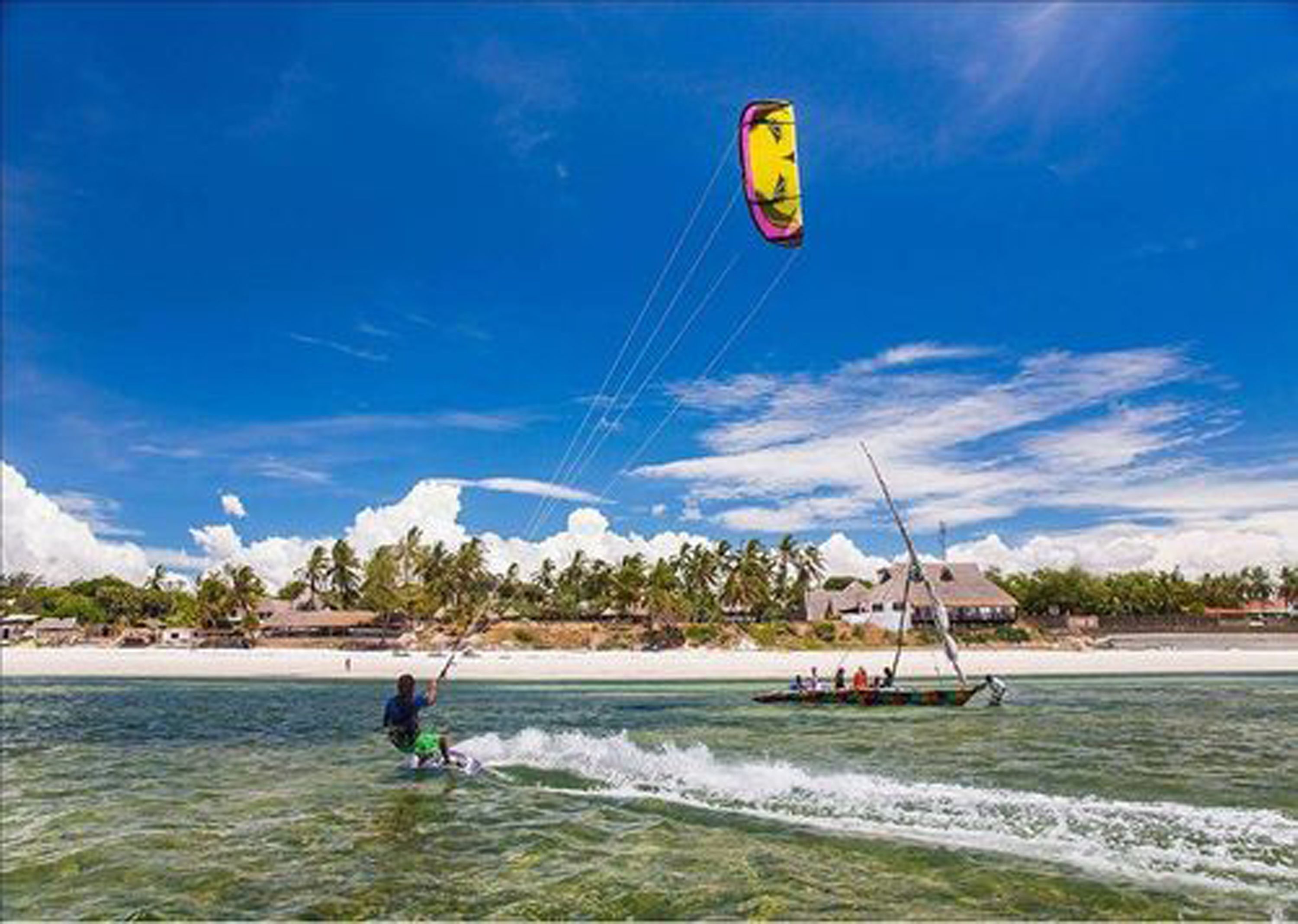 A beach with clear waters, a parasail, and a traditional boat with people on it.