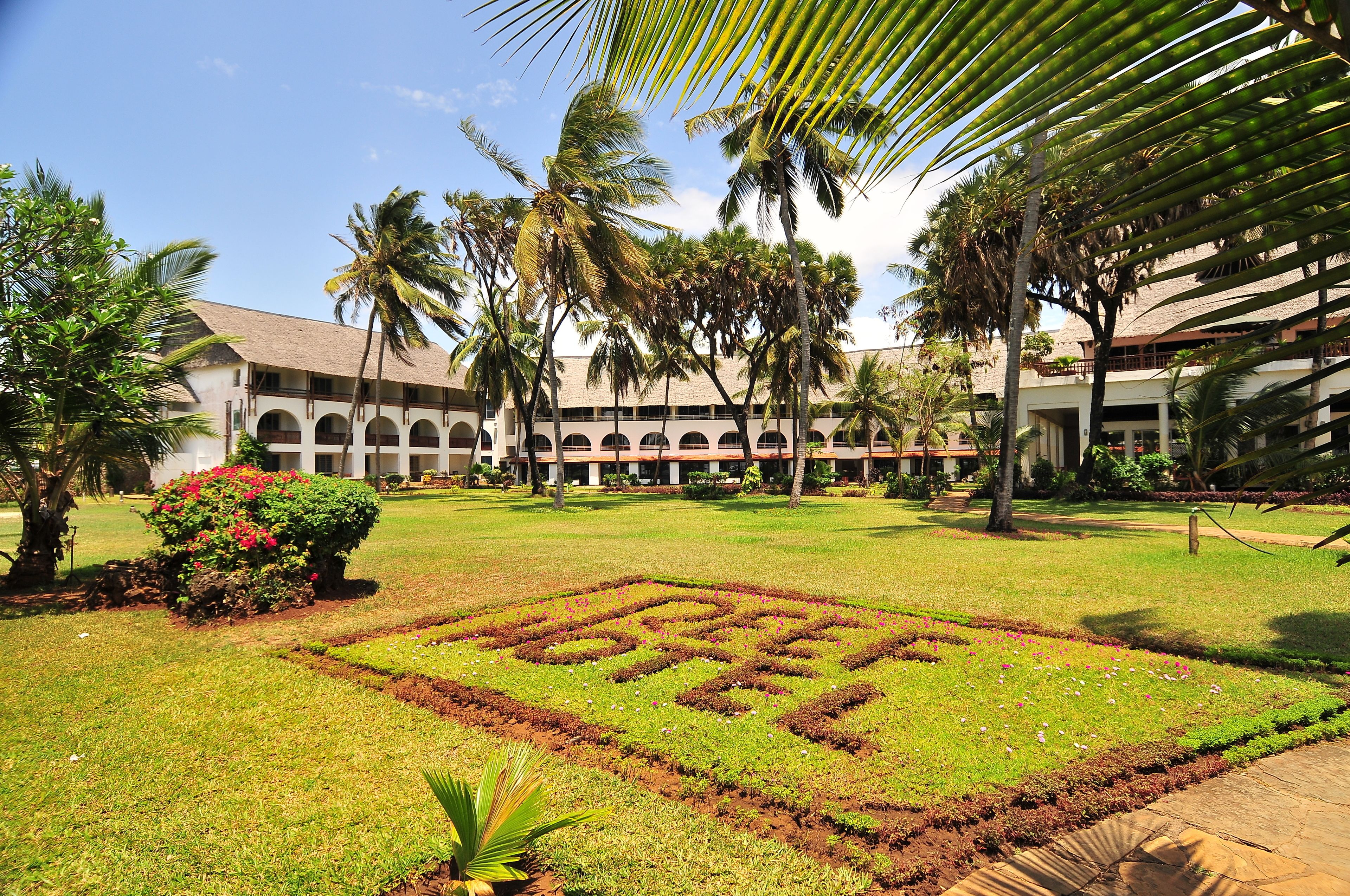 A courtyard with a geometric garden design, palm trees, and a building with arched walkways.