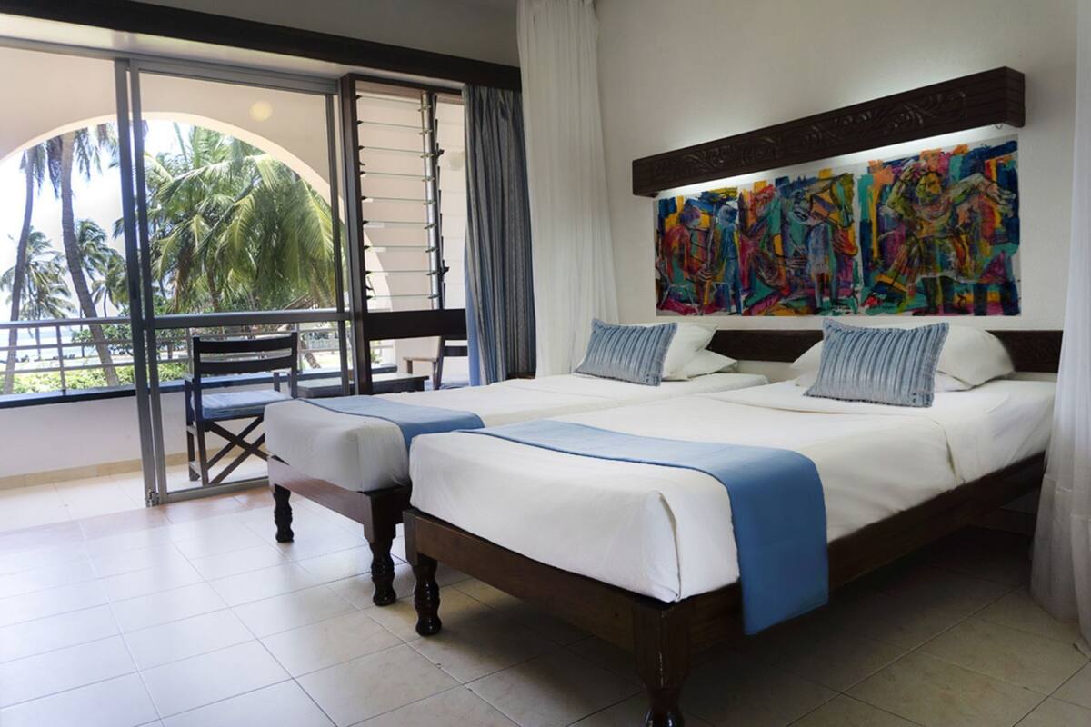 Habitación de hotel con dos camas, un balcón con una silla y un cuadro abstracto grande en la pared.