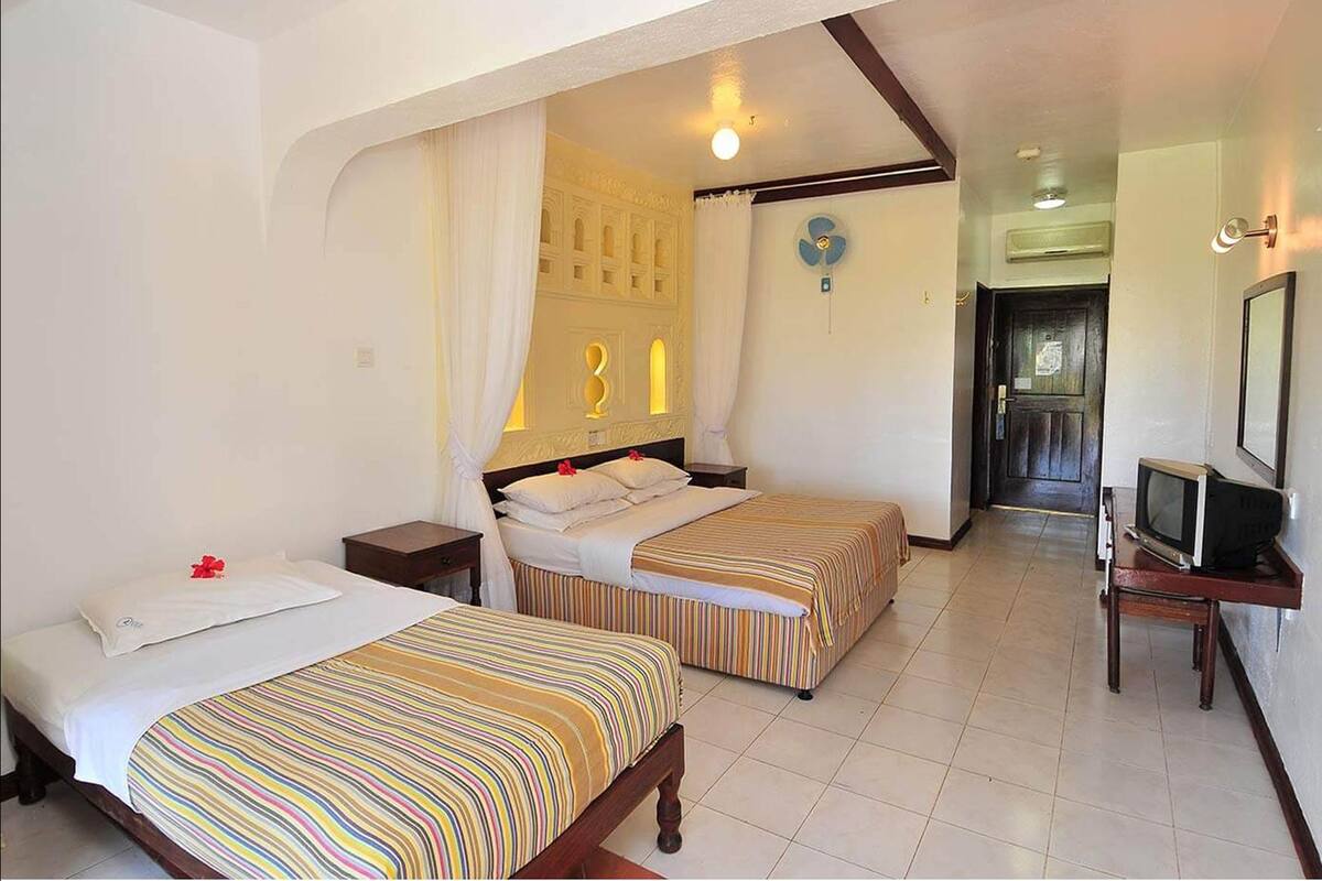 Habitación de hotel con dos camas, un televisor sobre un mueble bajo y una puerta que da a otra habitación.