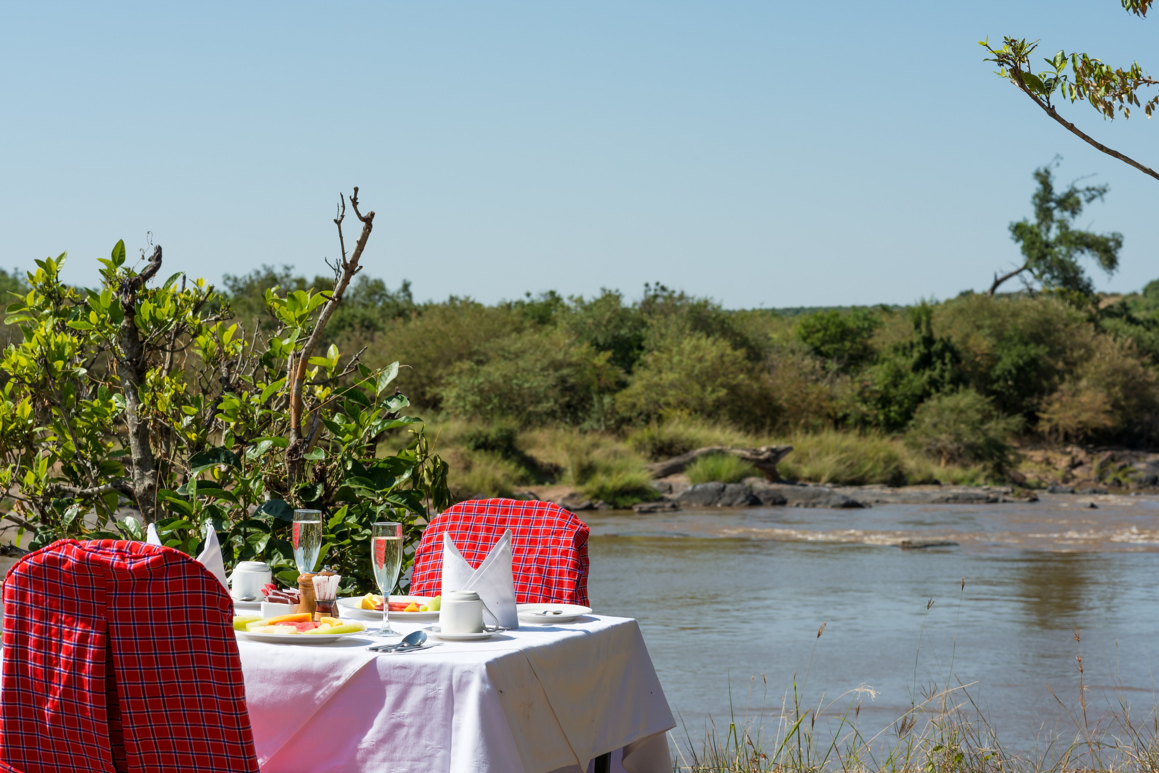 A dining table set up by a riverside with a view of the water and greenery.