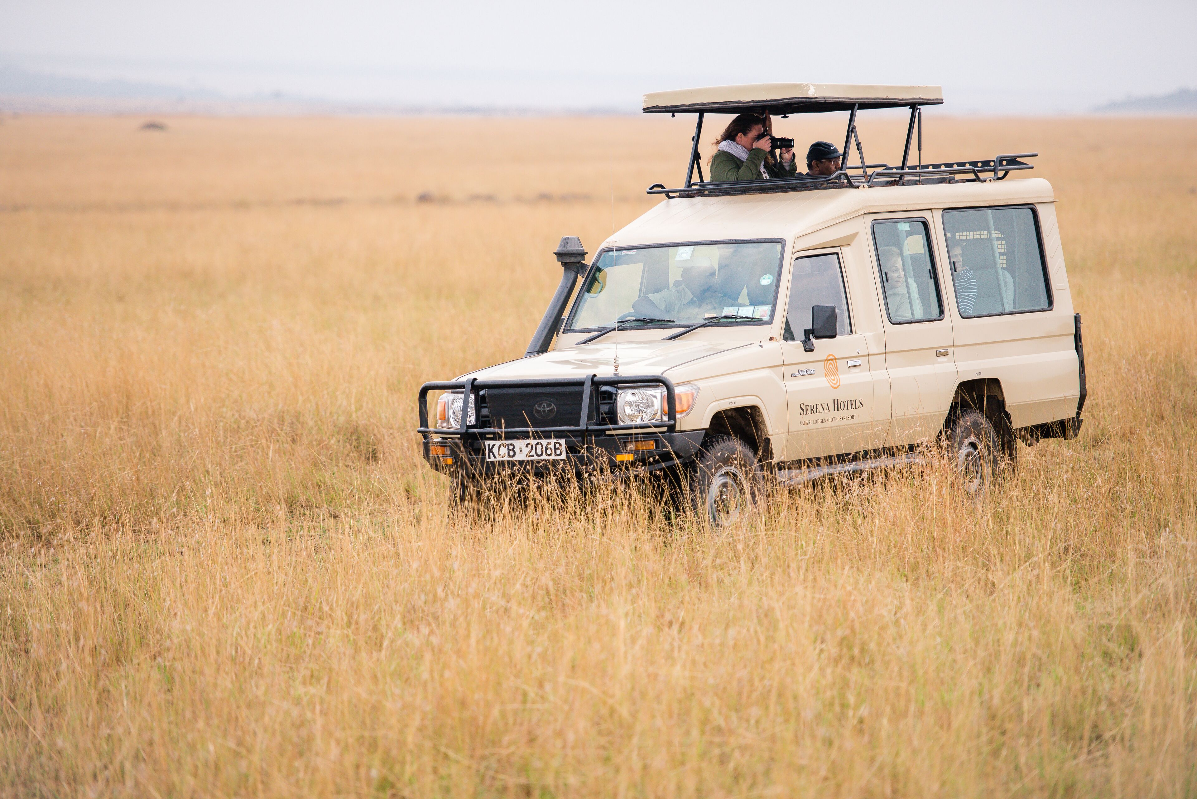 A safari vehicle with a roof rack and passengers, driving through a grassy field.