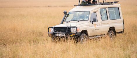 A safari vehicle with a roof rack and passengers, driving through a grassy field.
