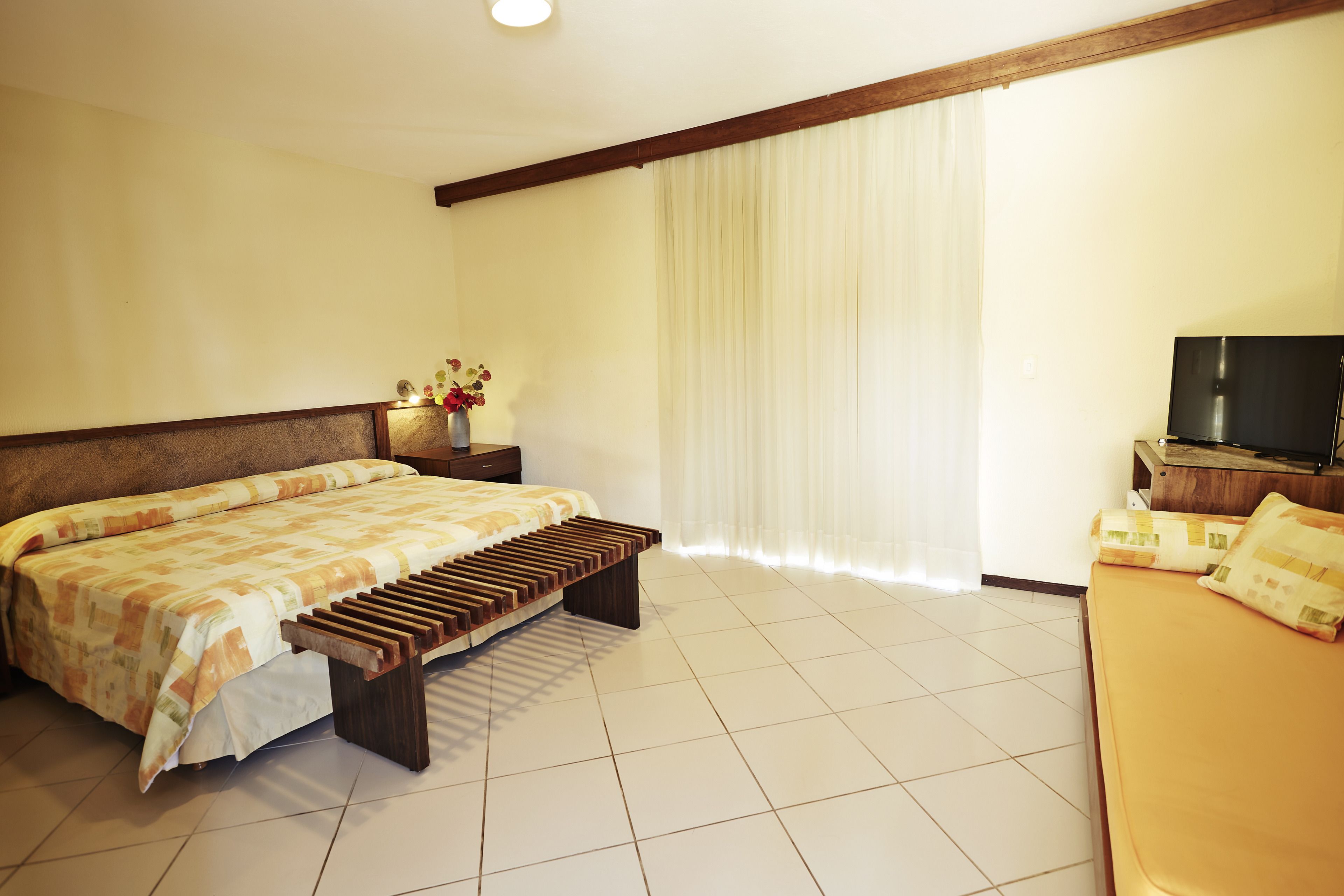 A hotel room with a bed, a bench, a TV, and a window with curtains.