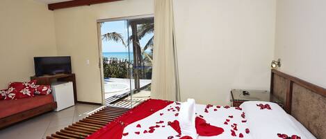 A hotel room with a bed adorned with rose petals and a white towel, a television, a small refrigerator, and a sofa with red and white pillows.