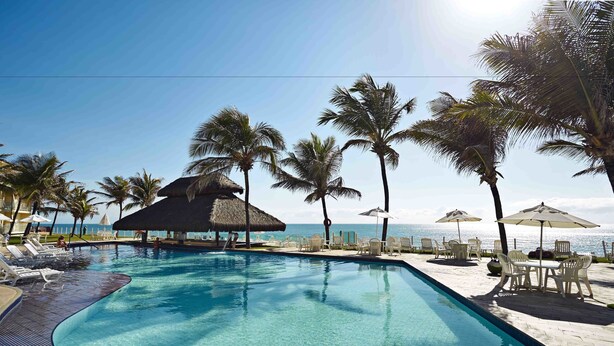 A clear swimming pool with lounge chairs and umbrellas, surrounded by palm trees and a view of the ocean. - EL ARAM NATAL MAR (Natal)