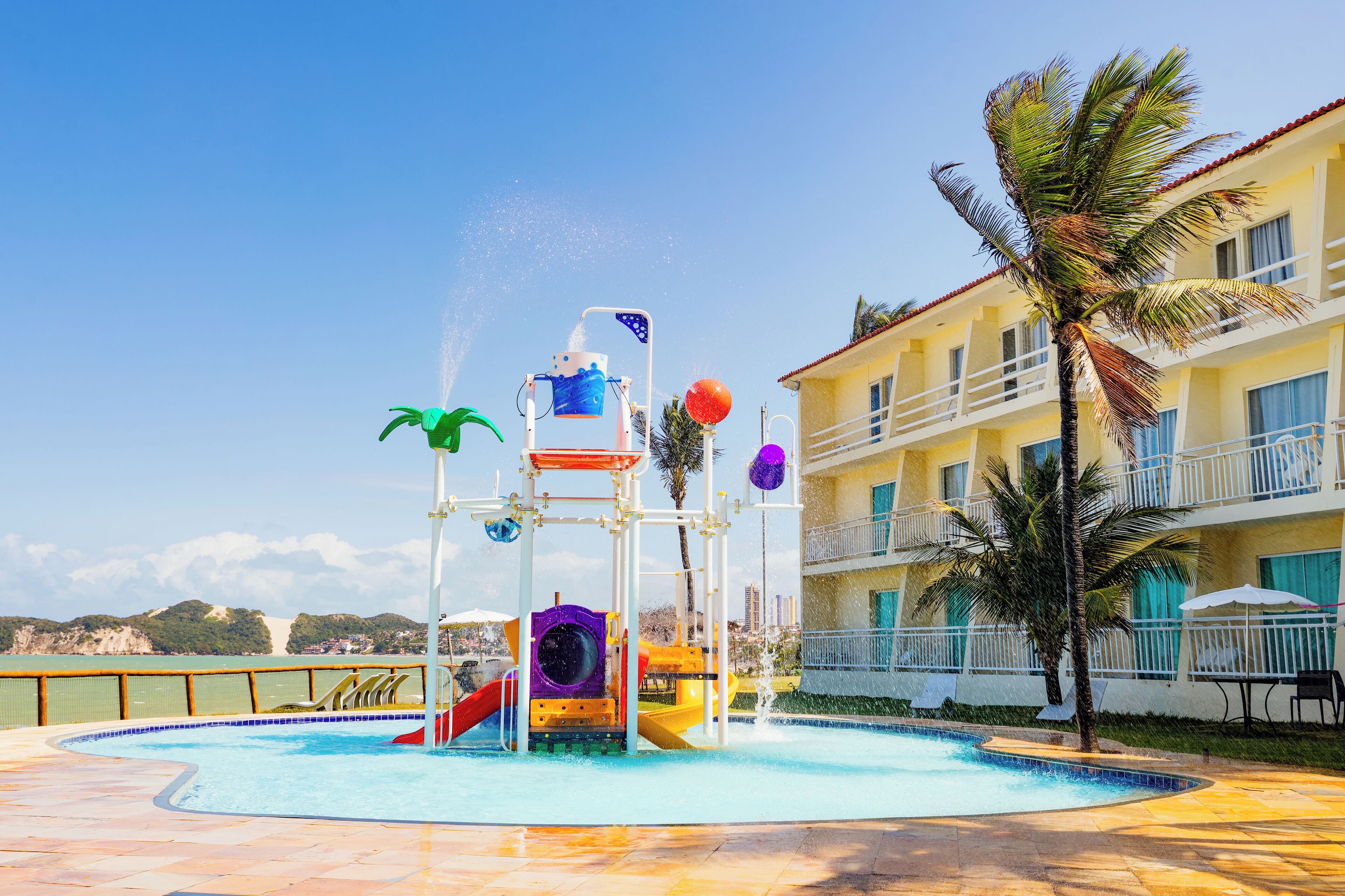 A water play area with slides and a pool, surrounded by a building with balconies and palm trees.