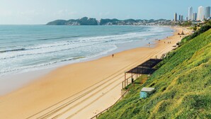 A beach with a sandy shore, a grassy hillside, and a city skyline in the background. - EL ARAM NATAL MAR (Natal)